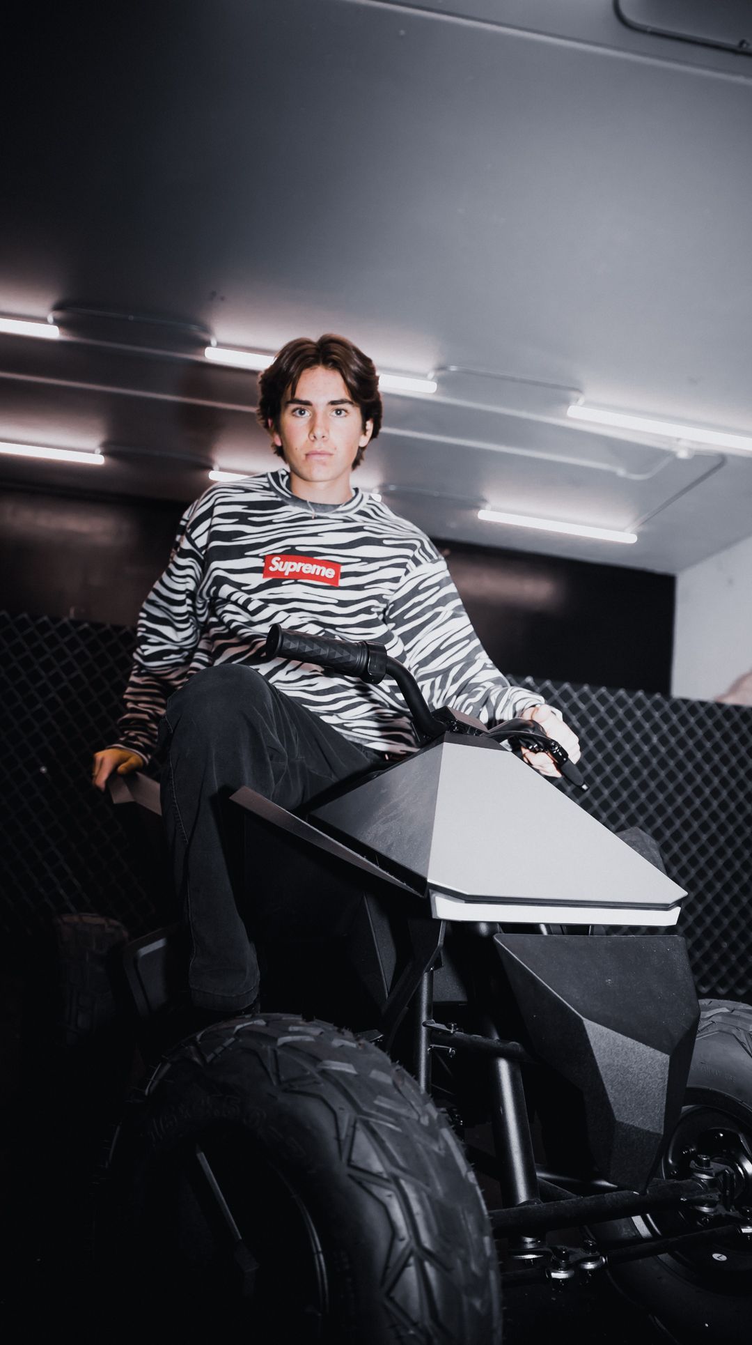 Fashion model in a zebra print sweatshirt poses on a futuristic vehicle in a dark studio setting.