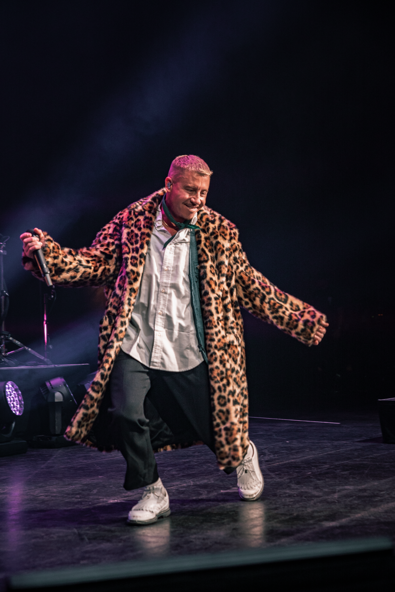 Performer wearing leopard print coat striking dynamic poses during a theatrical stage performance.