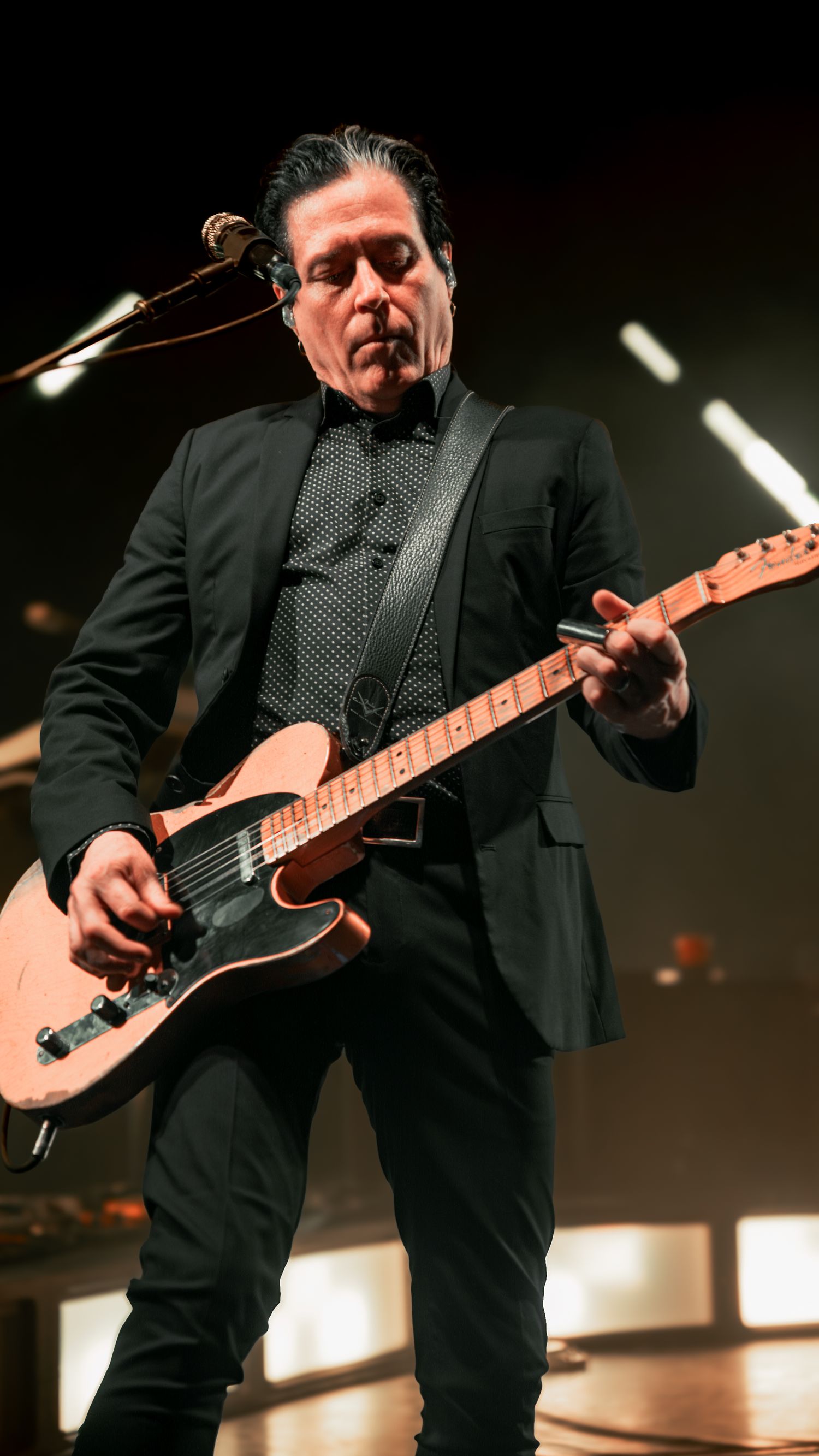 Guitarist in black suit plays electric guitar during dramatic performance on illuminated stage.