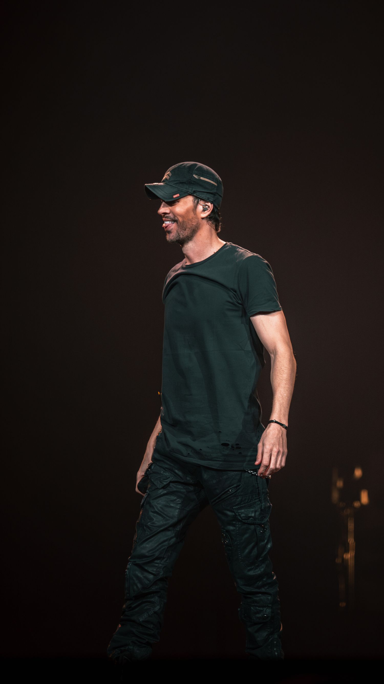 Performer in black t-shirt and cap smiling on dimly lit concert stage.