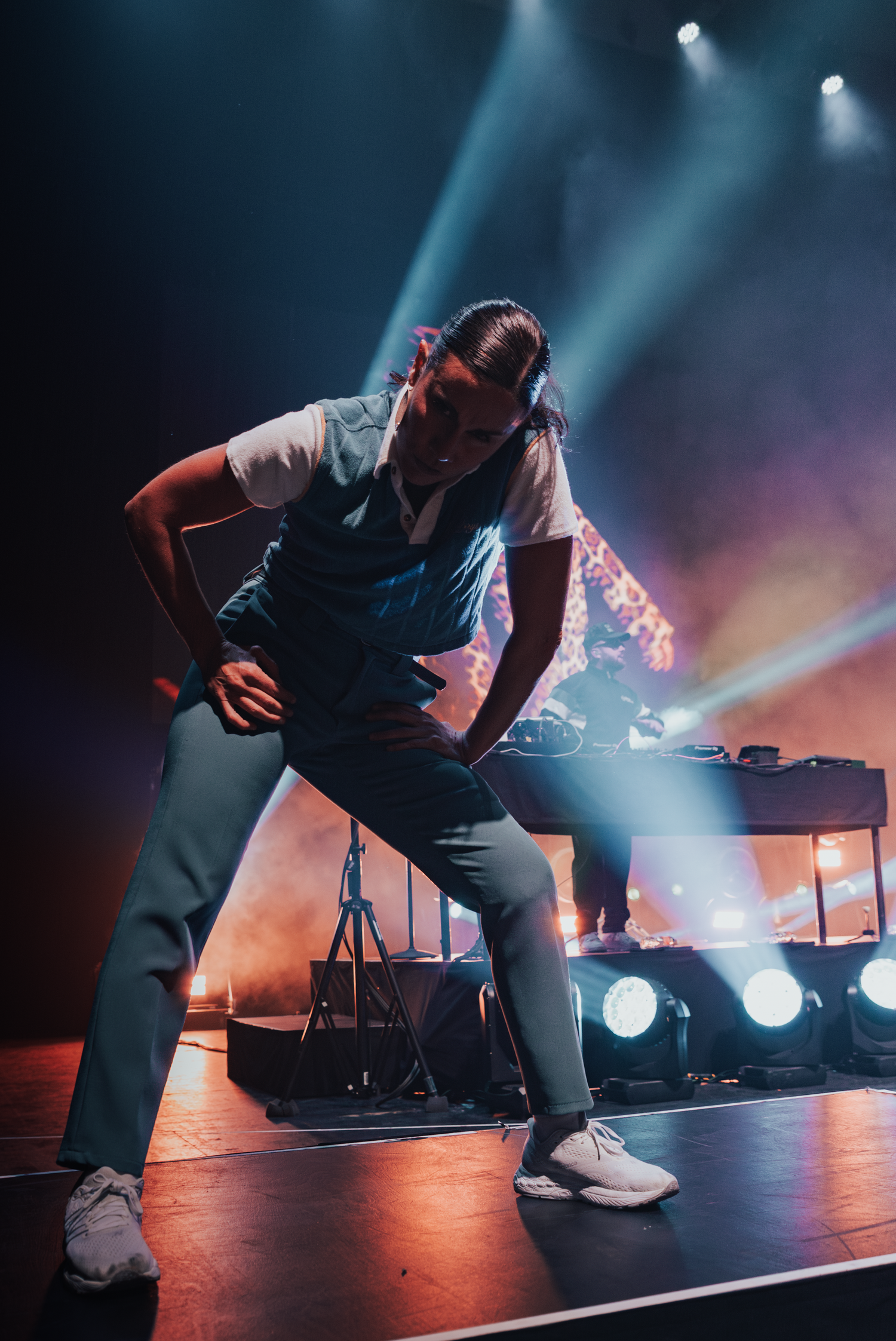 Dynamic stage performer in utility vest strikes an energetic pose under colorful concert lighting.