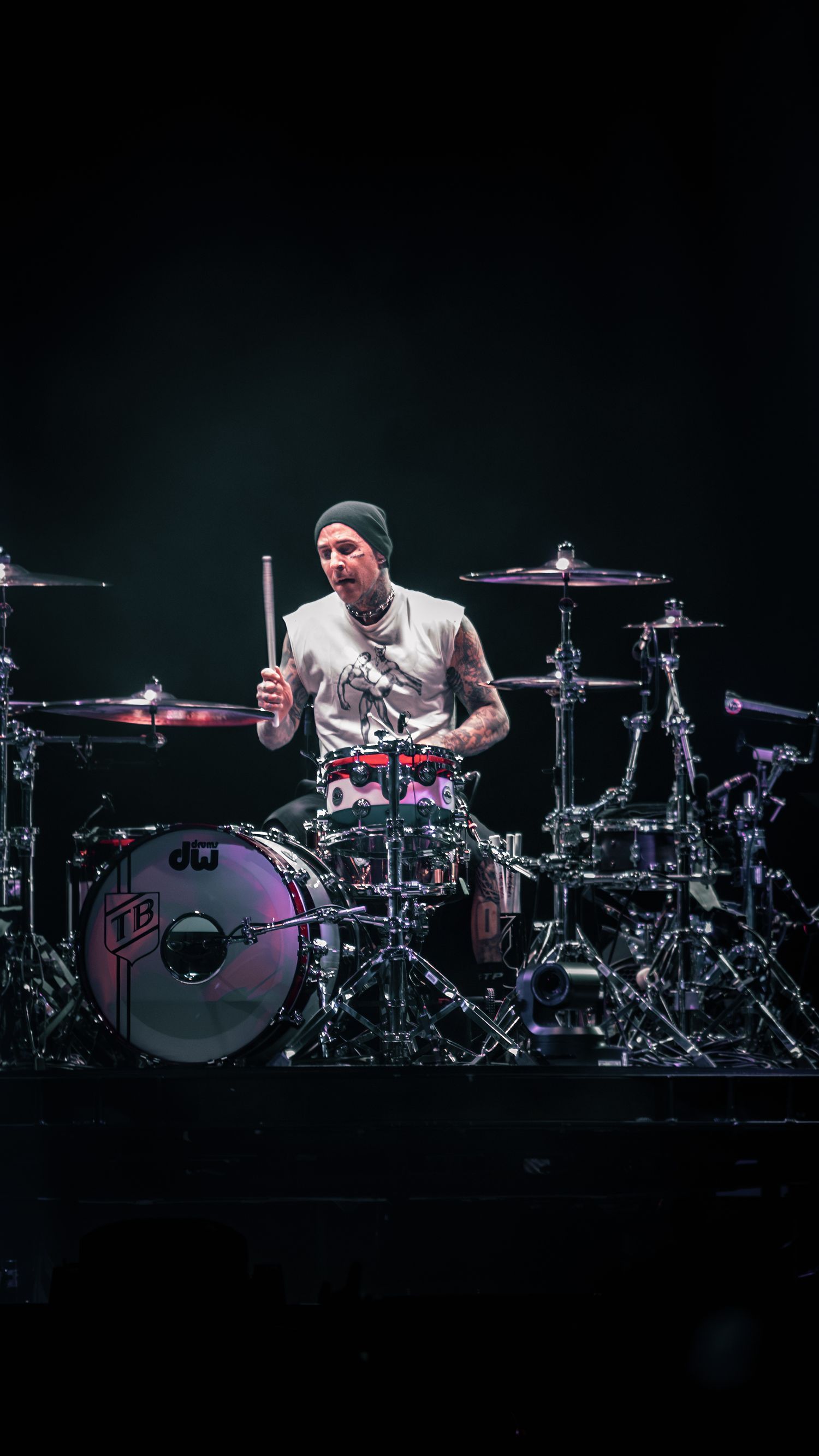 Drummer performs behind extensive drum kit setup in dramatic stage lighting.