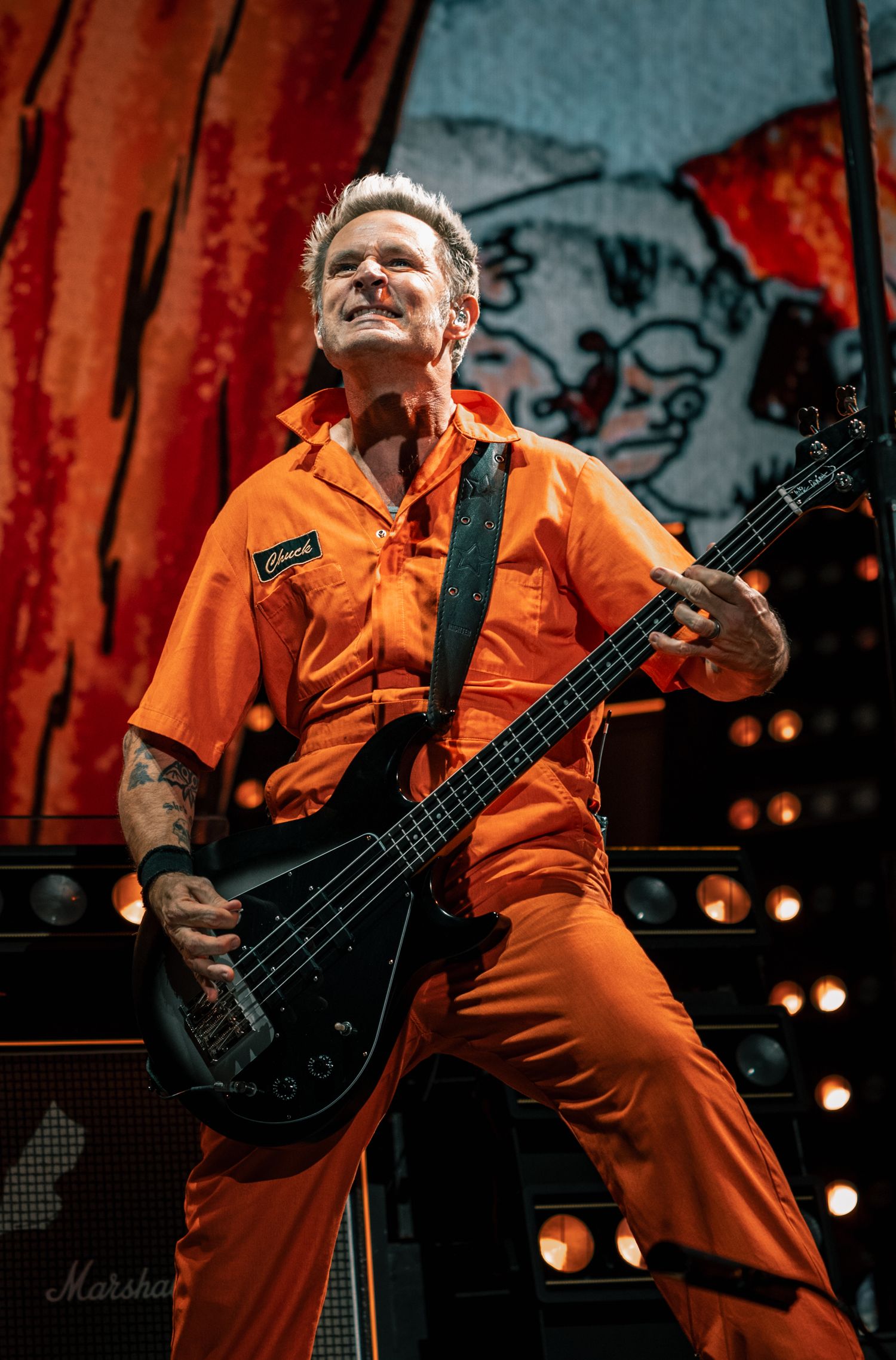 Bass player performs in bright orange jumpsuit against red backdrop on stage with Marshall amp.
