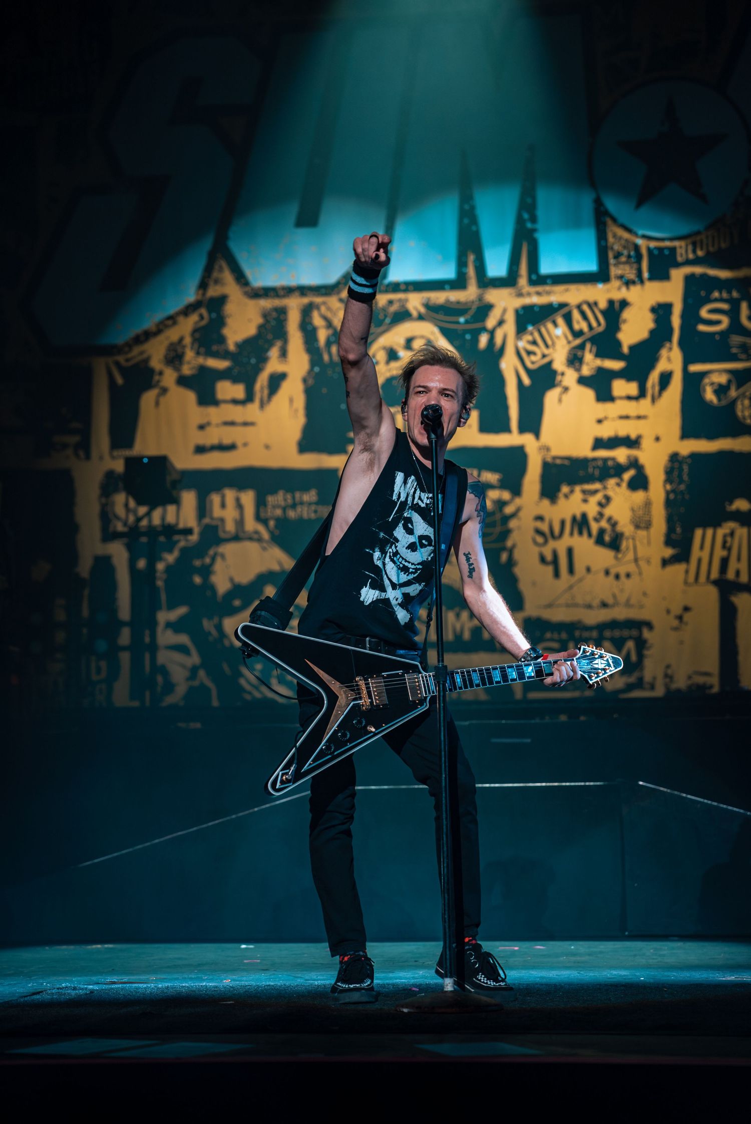 Performer with V-shaped guitar plays against graffiti backdrop under teal stage lighting.