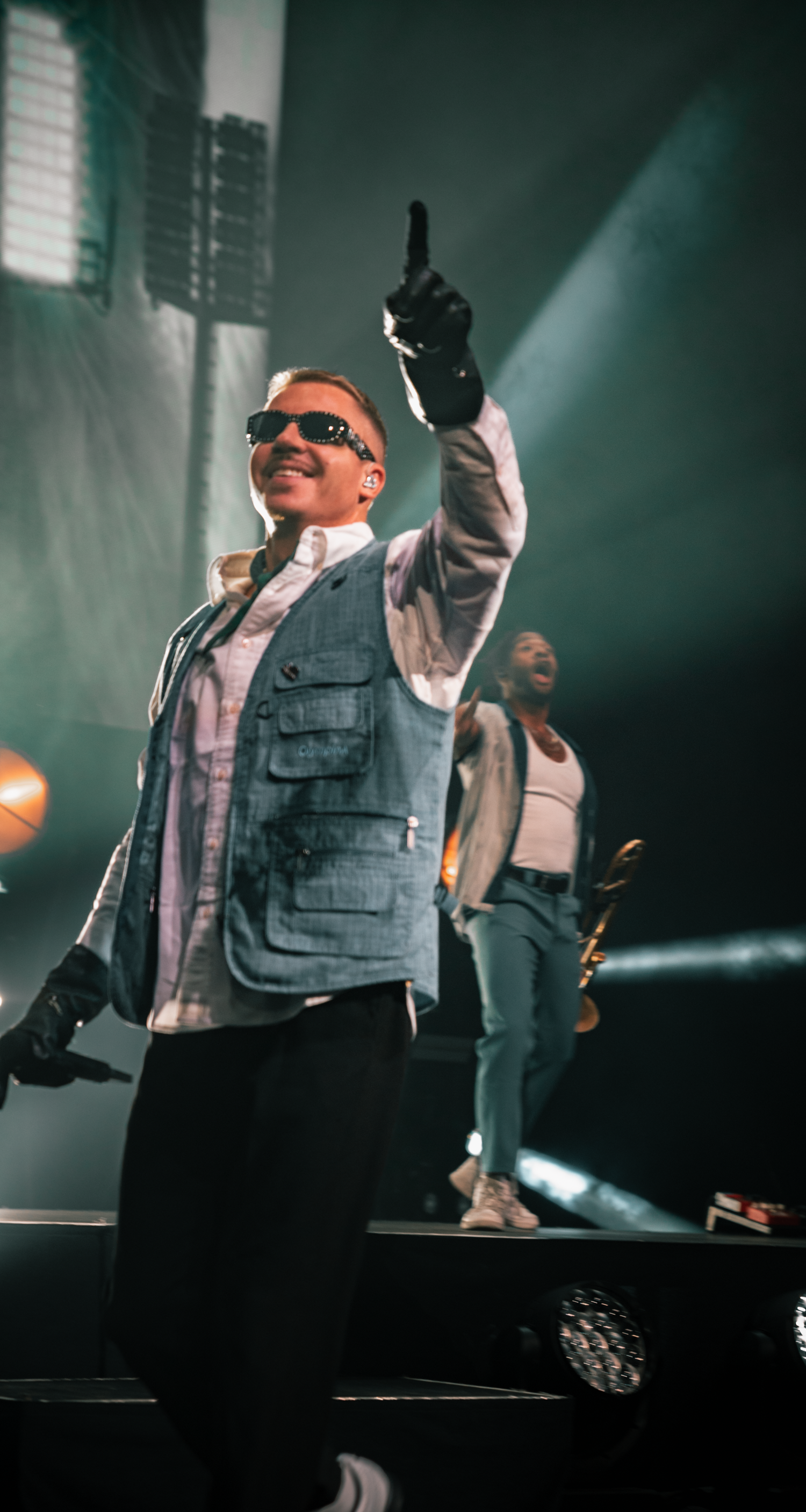 Concert performer in utility vest and sunglasses energetically moves on illuminated stage.