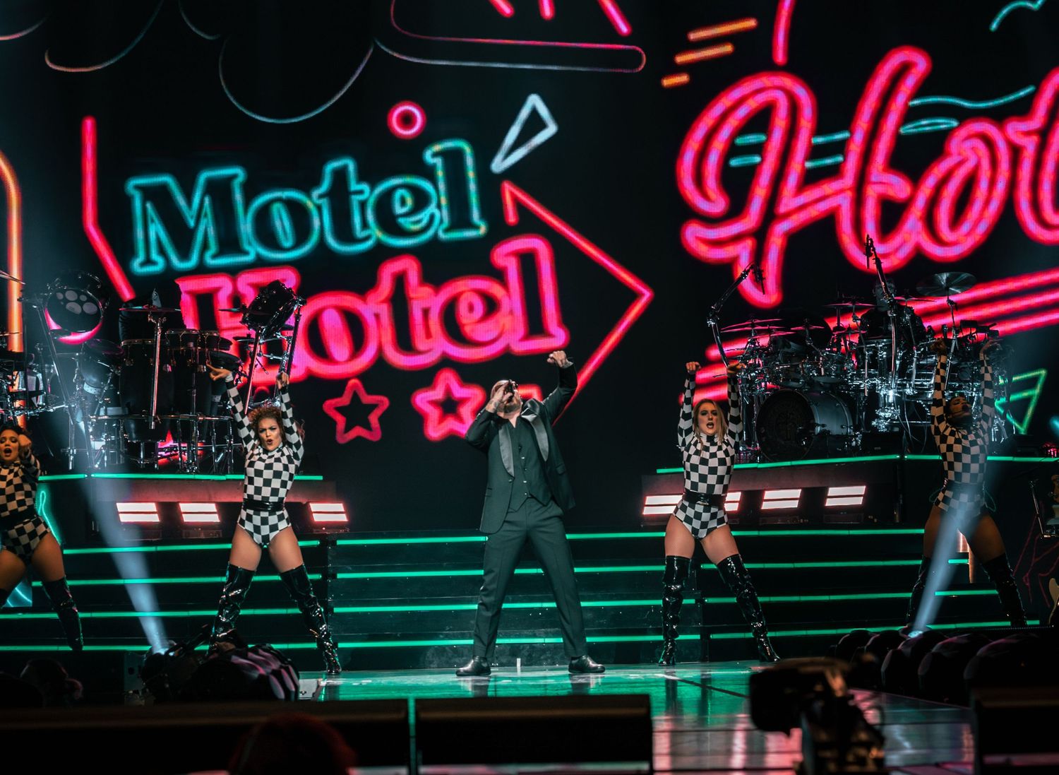 Concert stage with neon 'Motel' sign and performers in matching outfits performing choreographed routine.