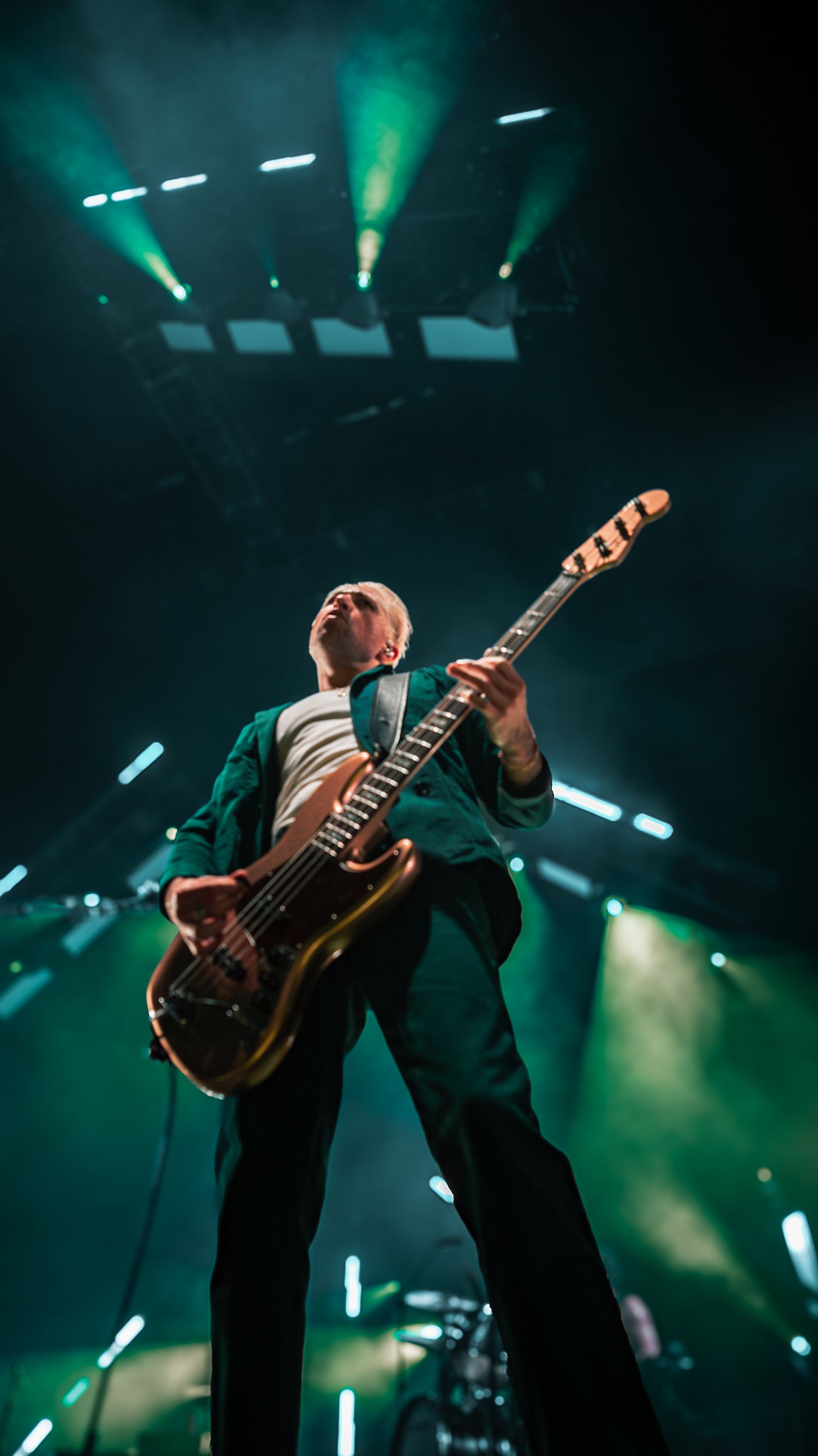 Performer plays guitar with dramatic stance against green concert lighting and smoke effects.