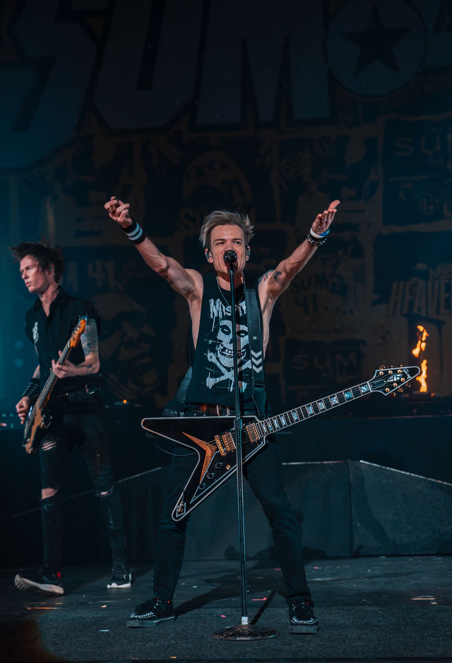 Musician plays Flying V guitar while posing with arms raised on dark concert stage.