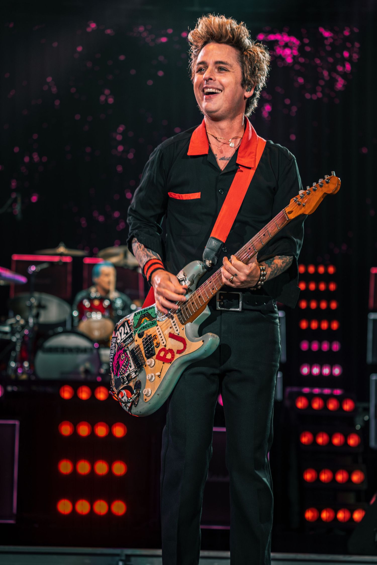 A guitarist performs with a decorated guitar on a concert stage with vibrant pink lighting and stage effects.