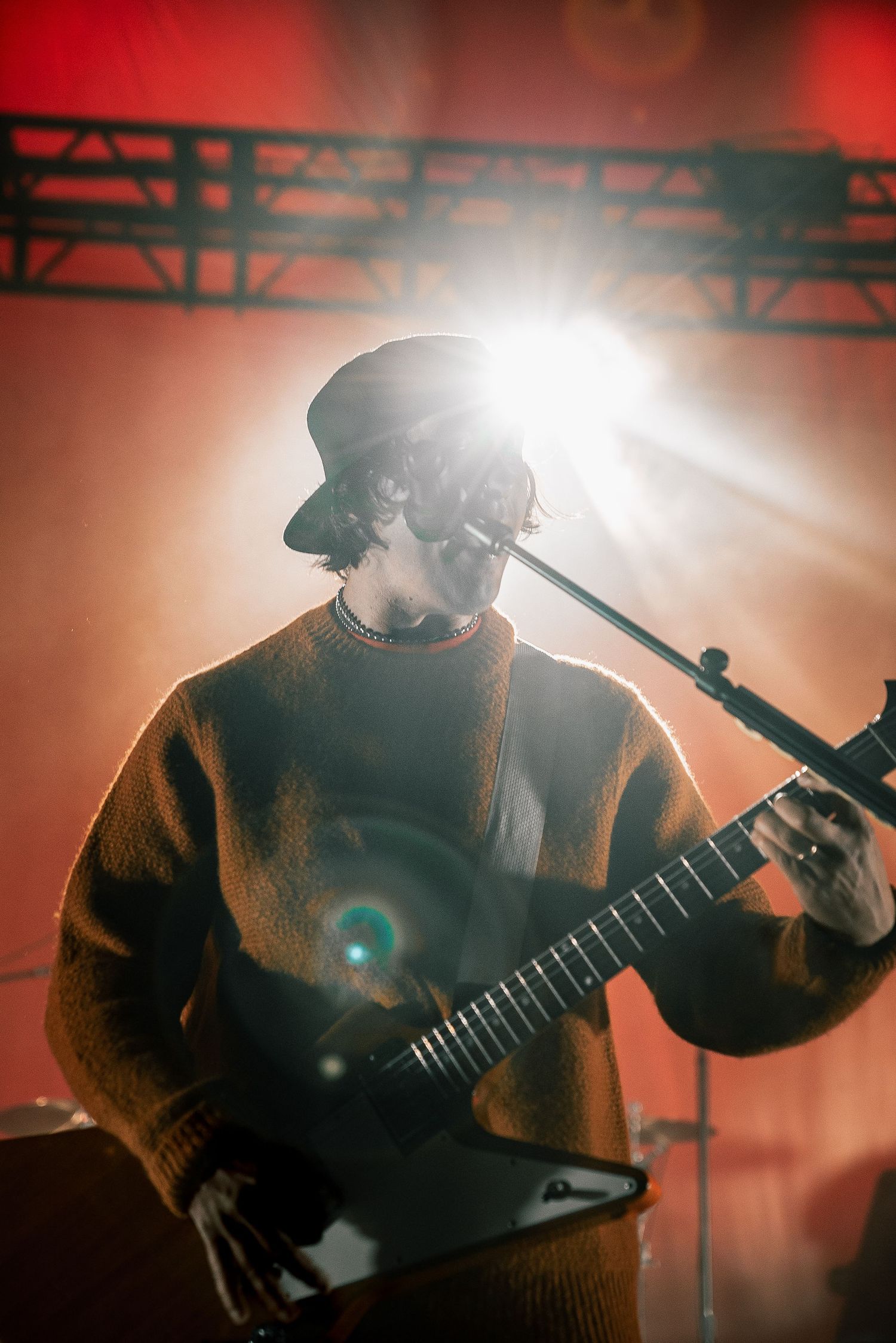 Performer in brown sweater plays guitar under bright stage lights creating atmospheric silhouette effect.