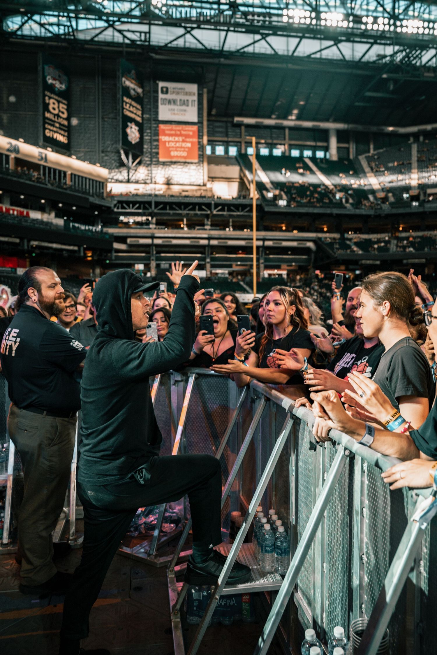Band members interact with excited fans across a barrier at indoor arena concert.