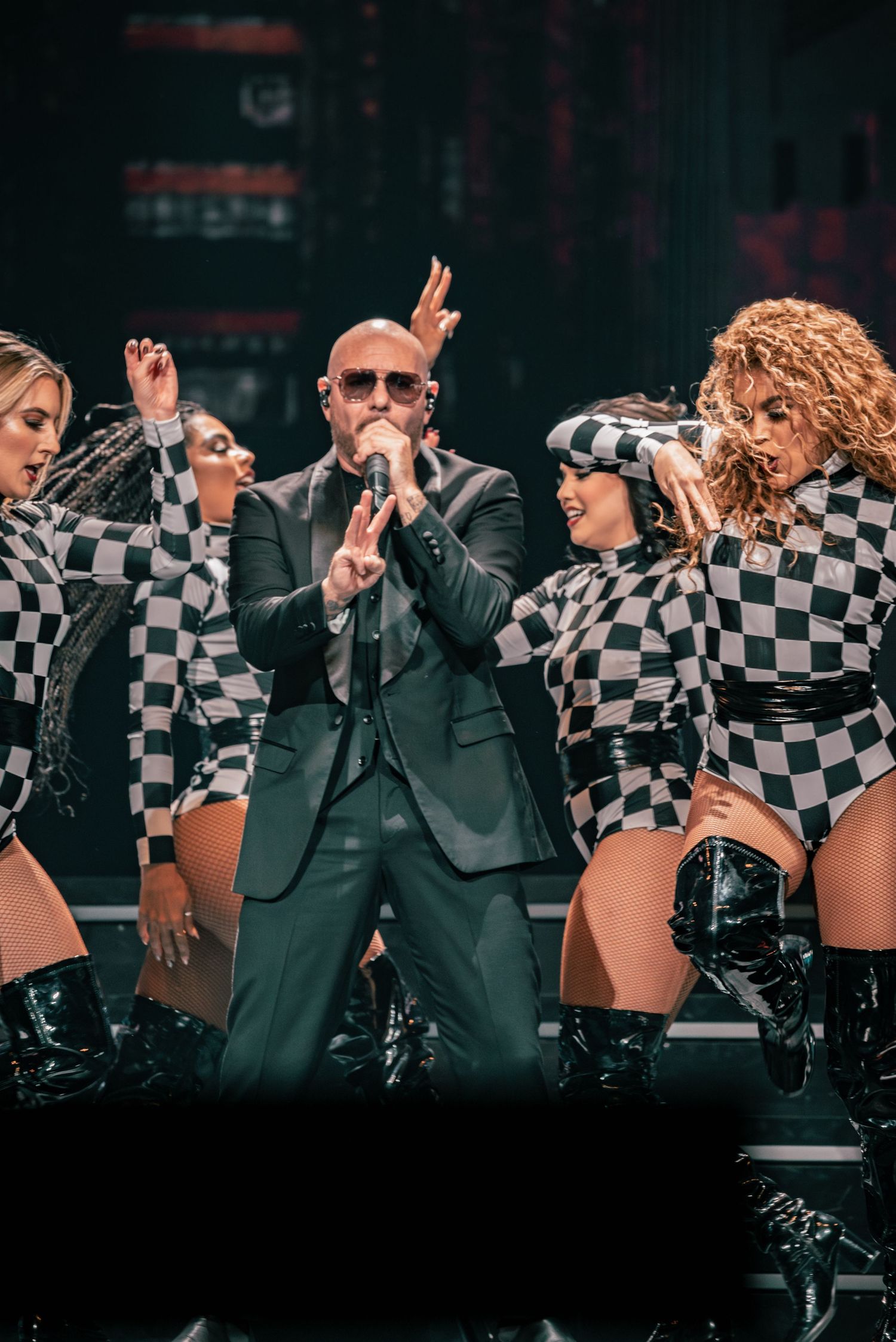 Energetic dance routine with performers in matching checkered outfits and suits on dark stage.