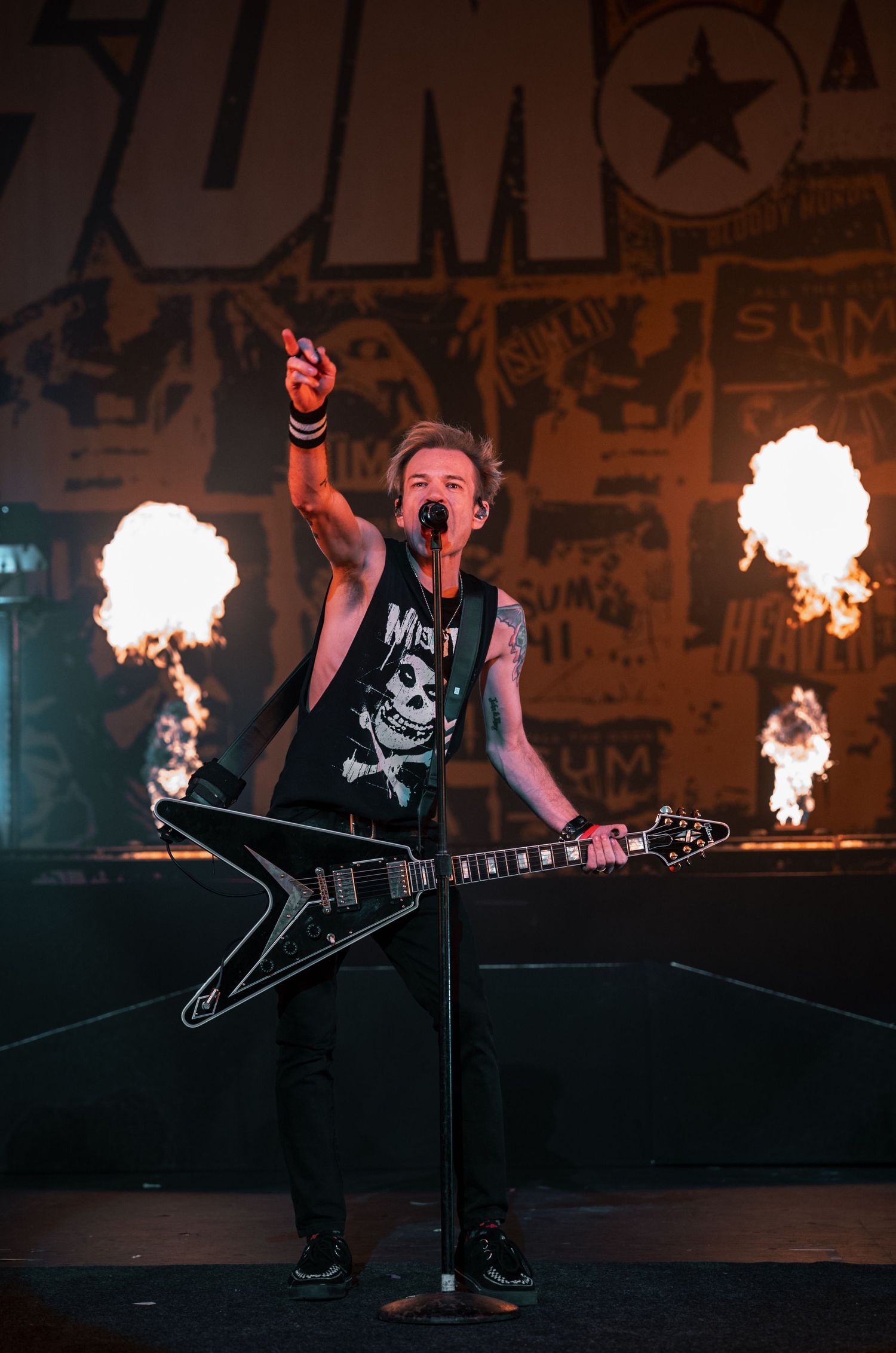 Musician plays flying V guitar on stage with pyrotechnic fire effects and graffiti backdrop.