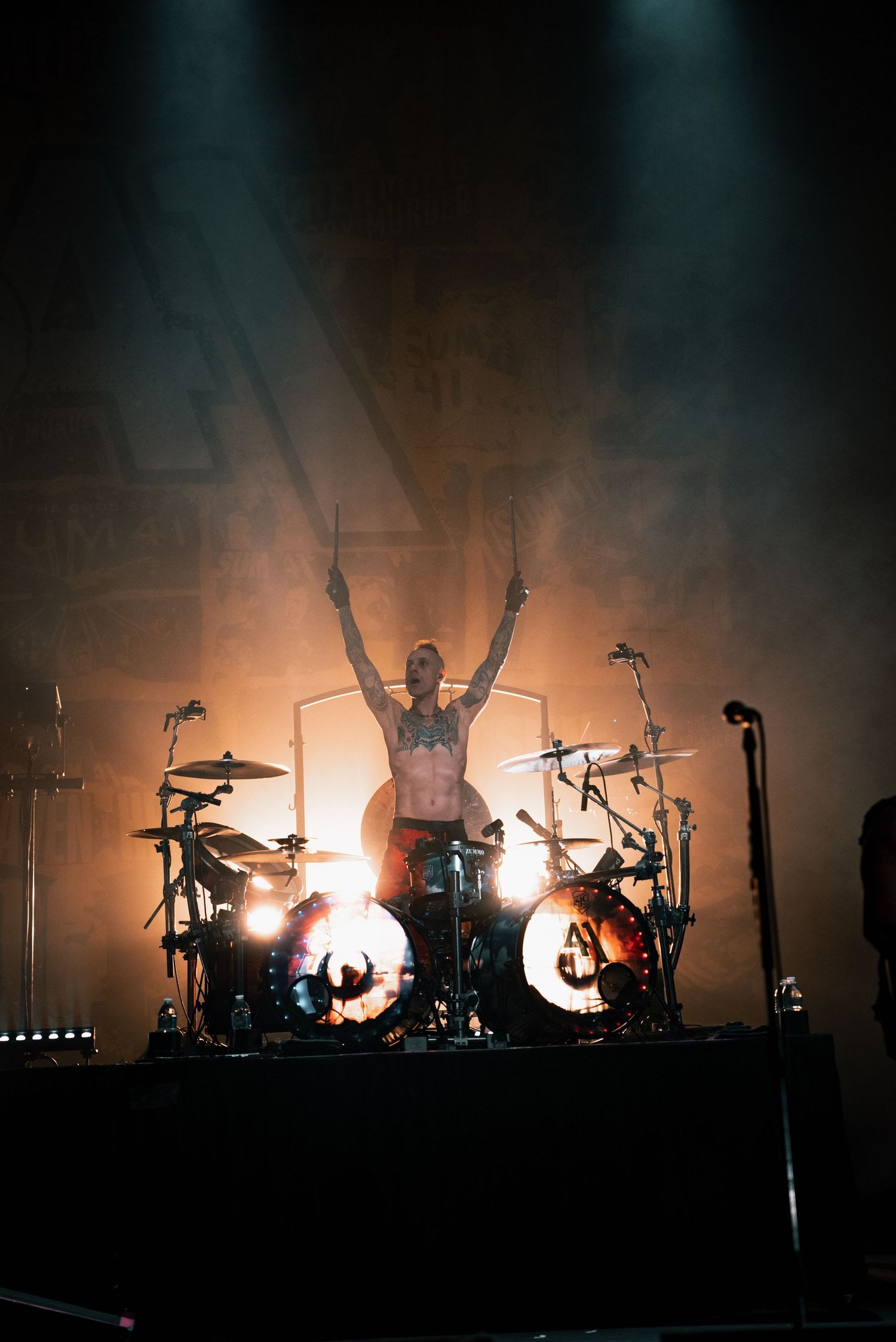 Silhouetted drummer raises arms triumphantly behind illuminated drum kit on concert stage.