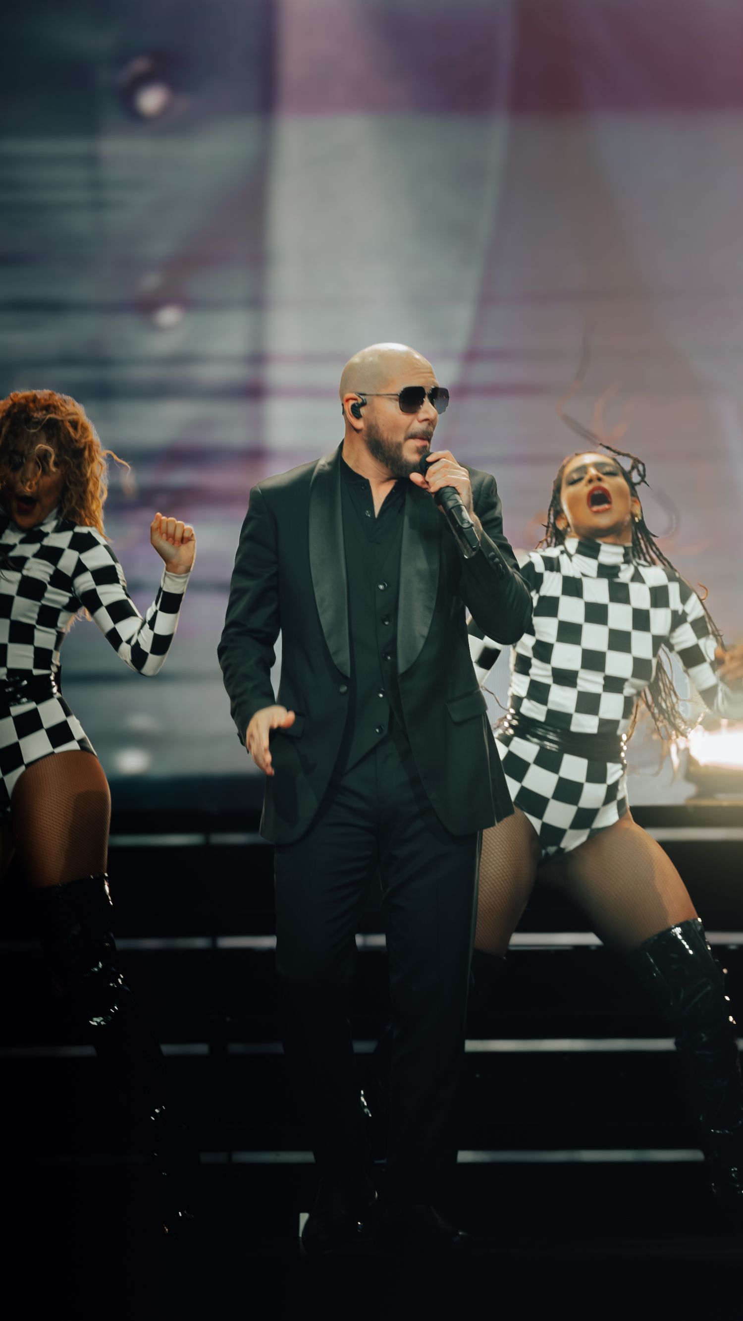 Concert performance featuring dancers in checkered bodysuits and tall boots alongside performer in dark suit.