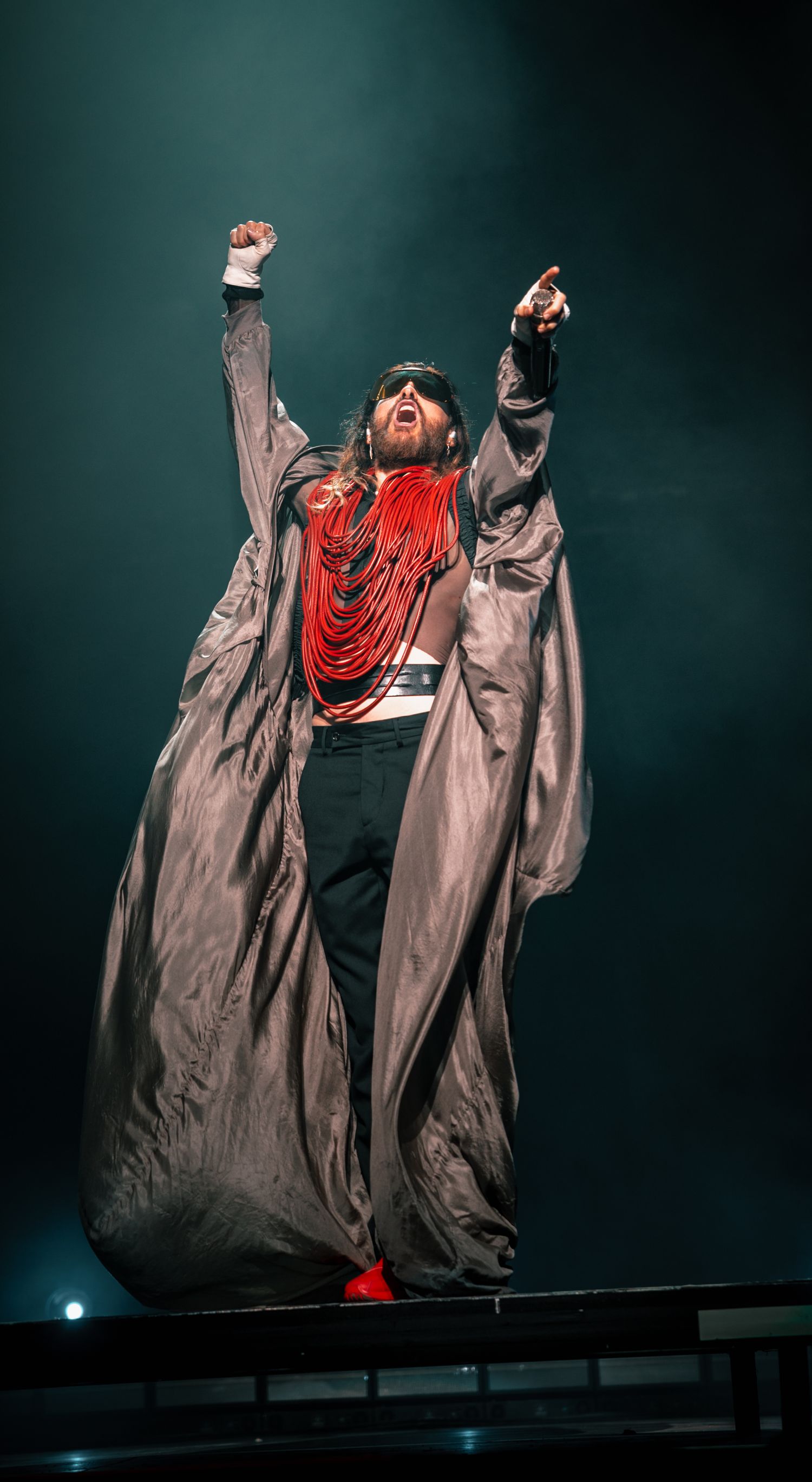 Performer in dramatic gray coat and red accent strikes powerful pose on elevated stage.