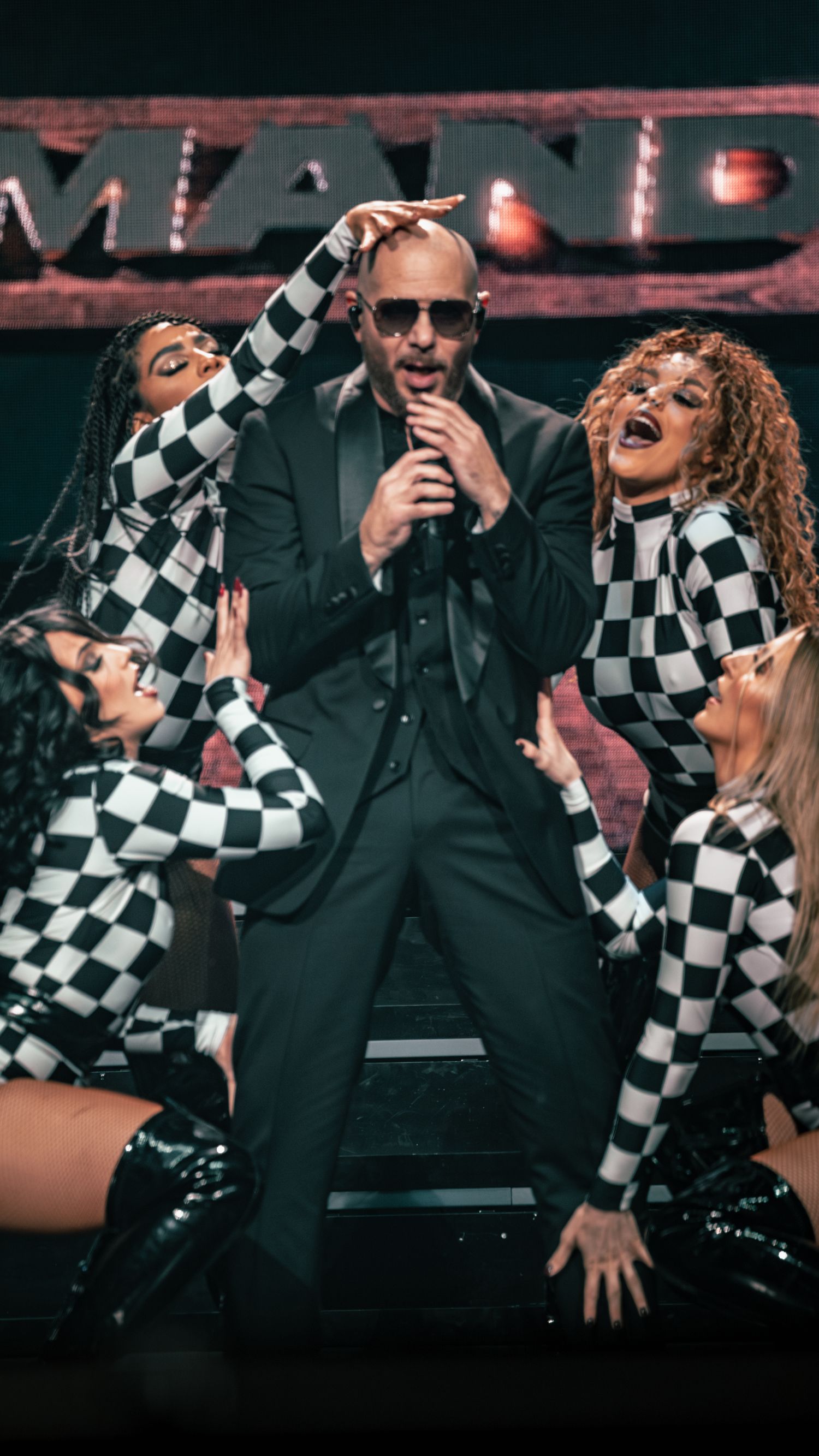 Concert scene with backup dancers in checkered bodysuits performing alongside suited singer on stage.
