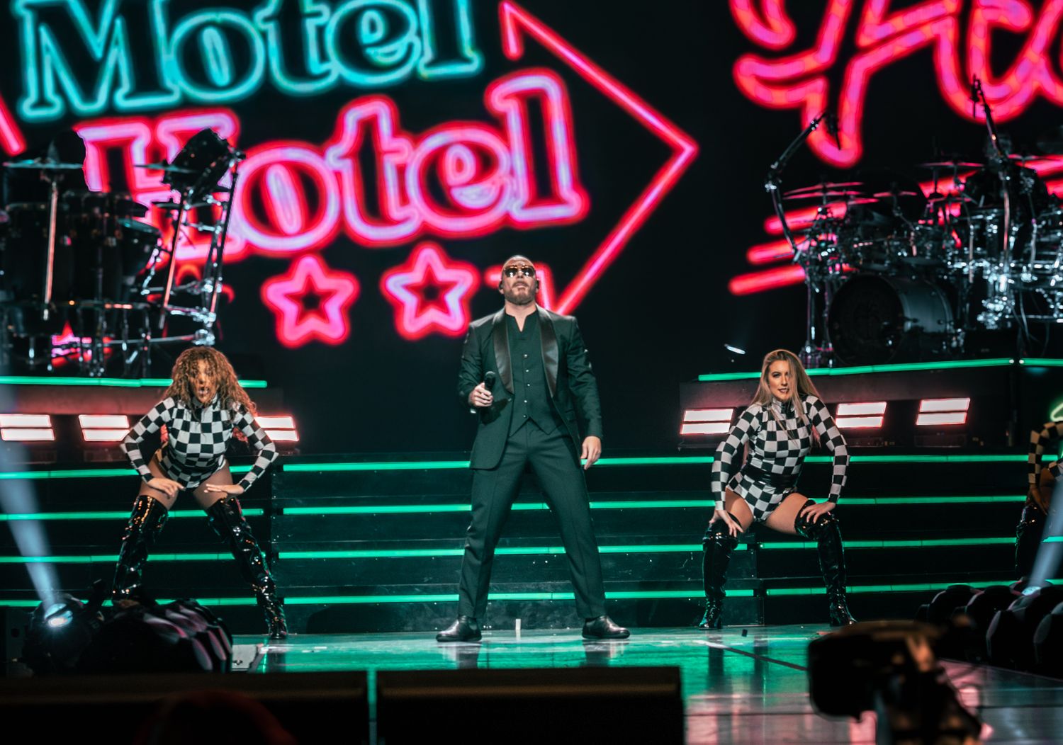 Live performance with neon motel sign backdrop and performers on stepped stage platform.