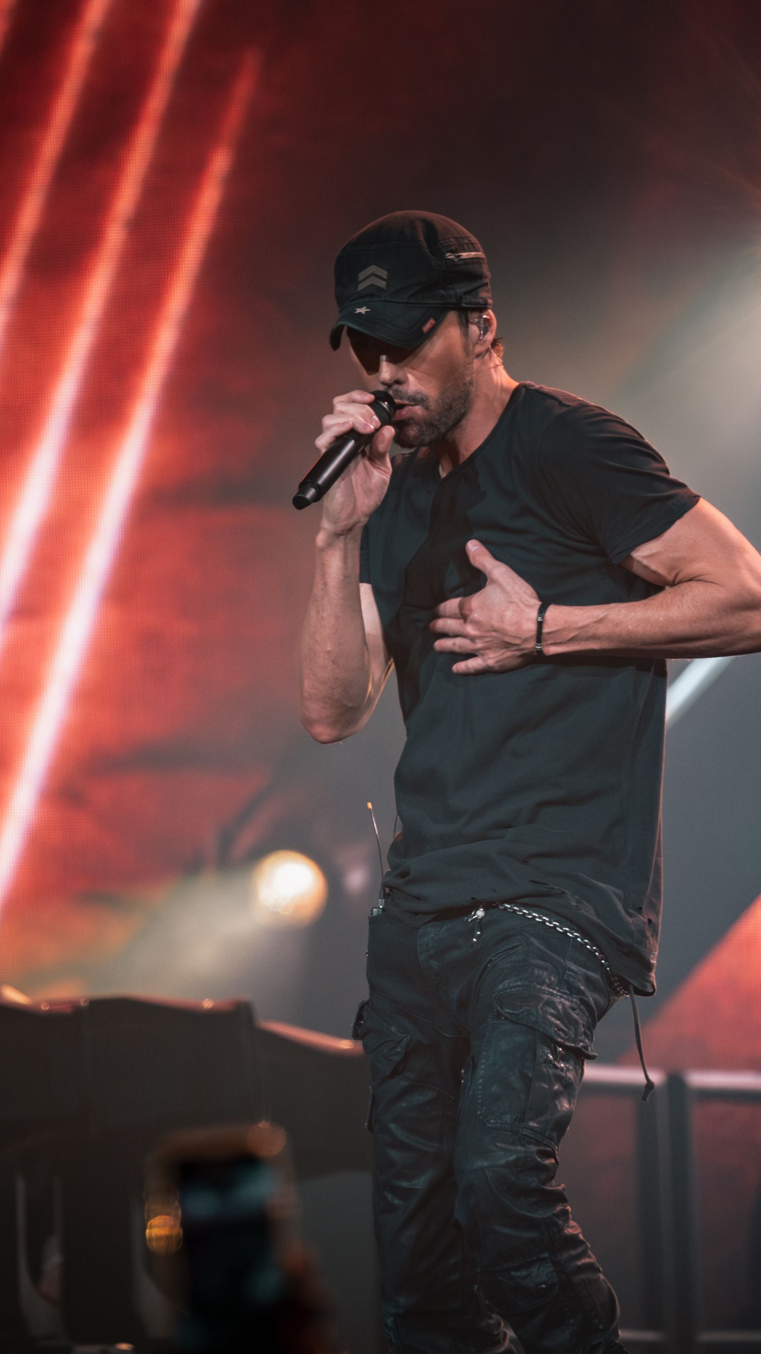 Concert performer wearing dark attire sings passionately into microphone against red stage lighting.