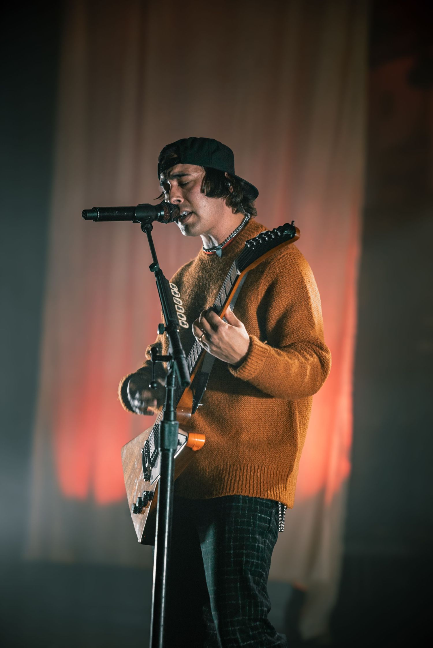 Stage performer in orange sweater sings into microphone with dramatic lighting effects.