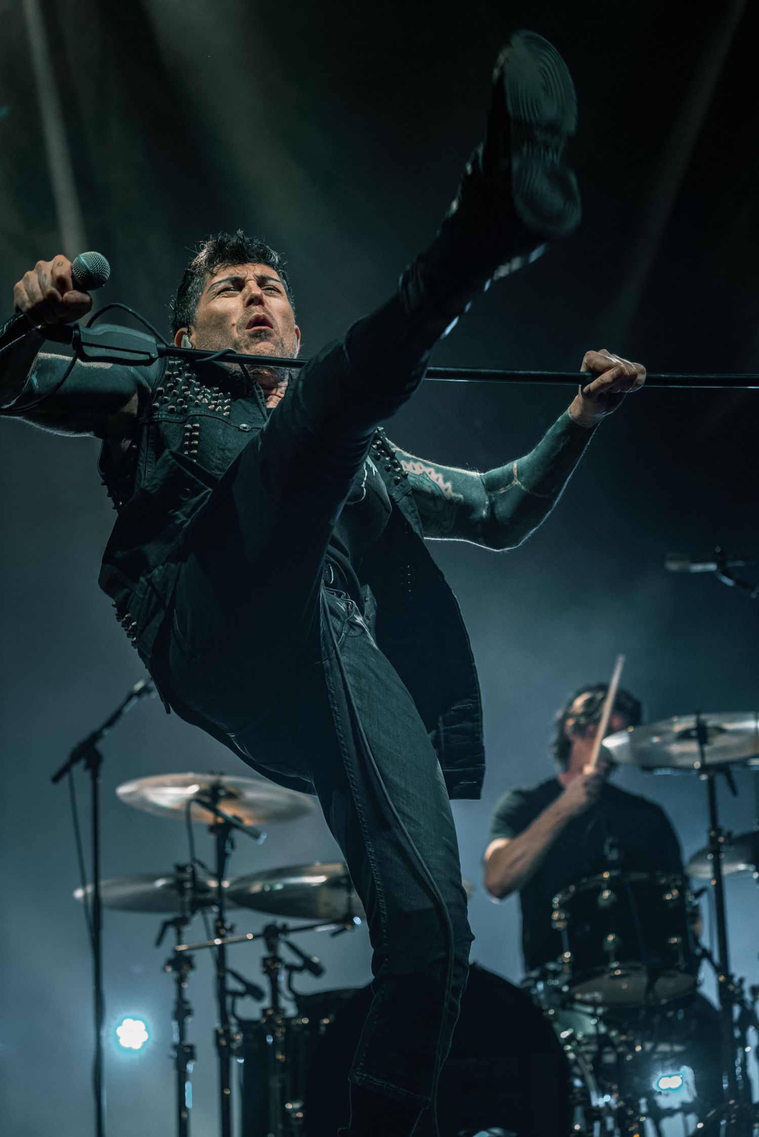 Performer leaping energetically on stage with drums and equipment visible in background.