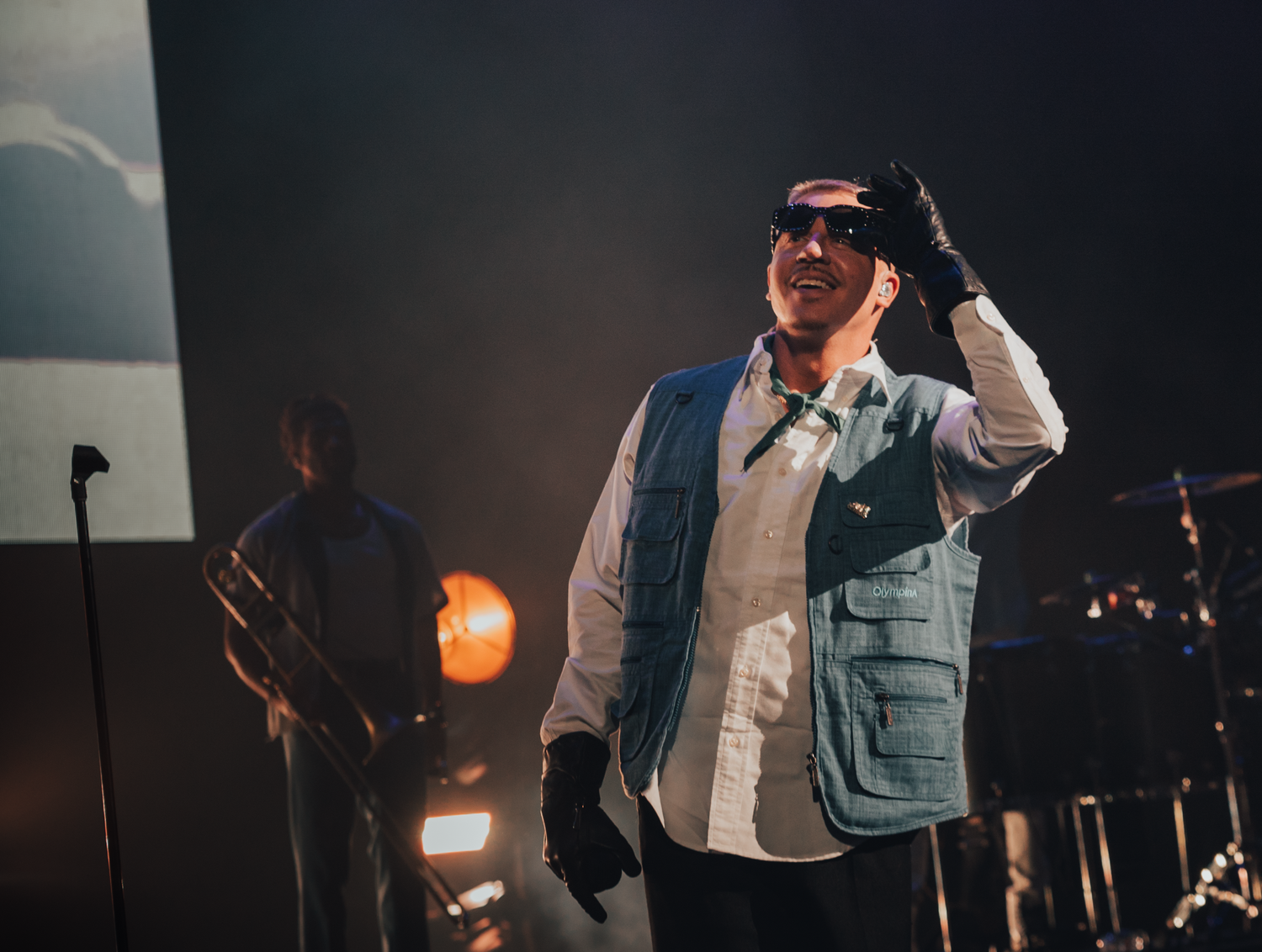 Performer in a white shirt and utility vest raises their hand during a dimly lit stage performance.
