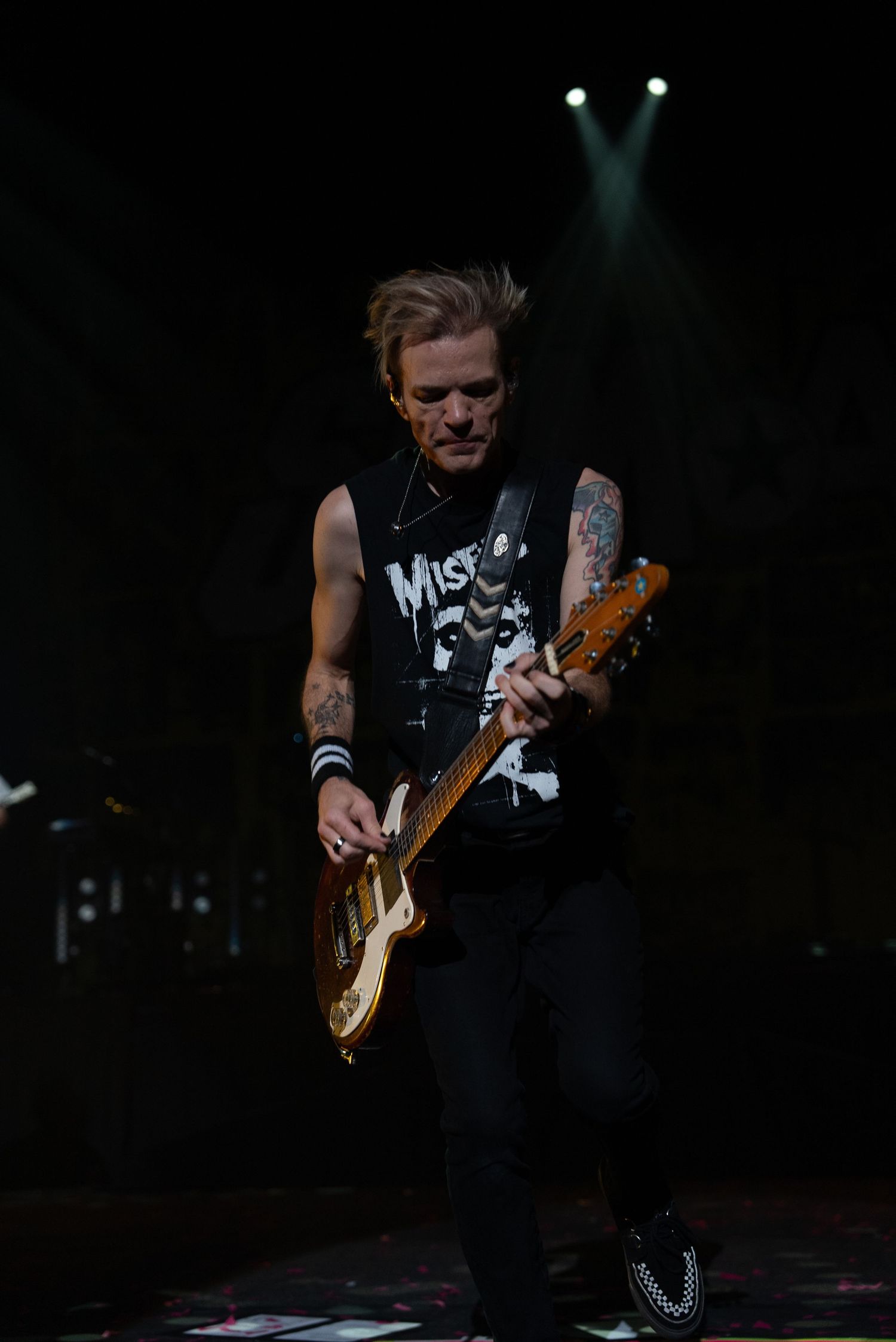 Guitarist in black sleeveless shirt performs under spotlight on dark stage.