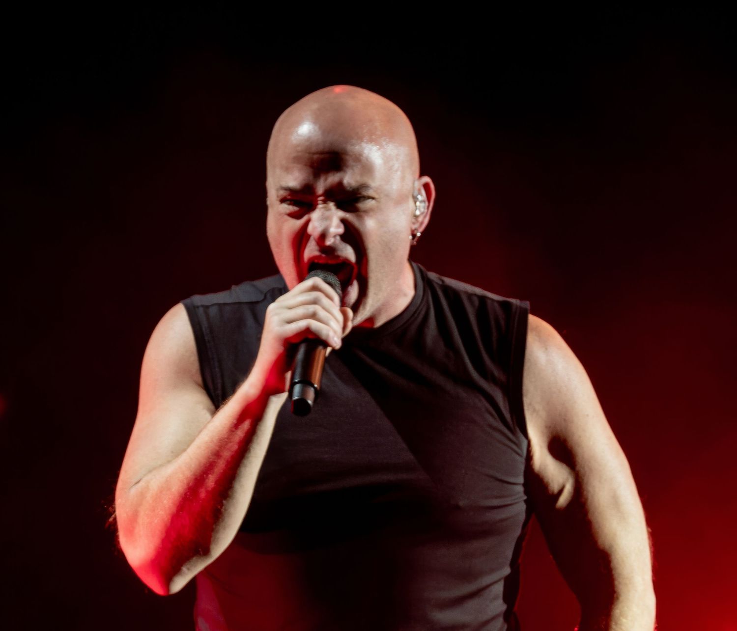 Performer in black tank top singing passionately on stage with red lighting effects.