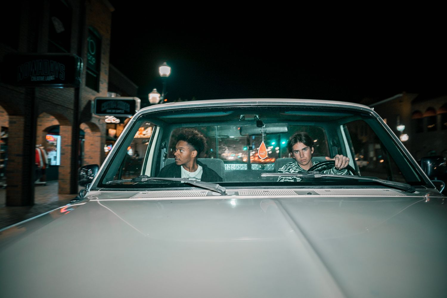 View through windshield of people in vintage car at night with city lights in background.