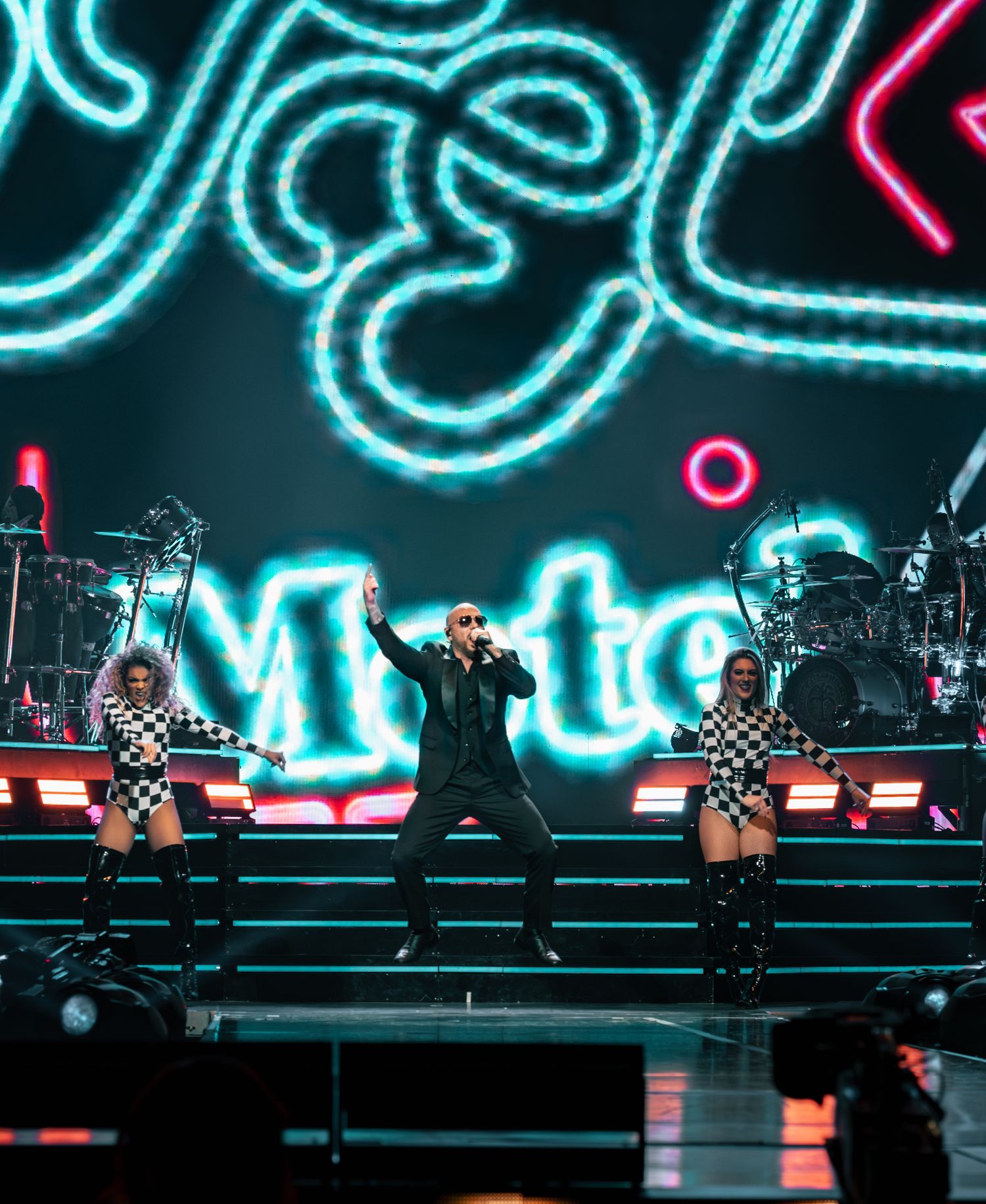Concert performer in black outfit with dancers in checkered outfits on stage with bright neon lights and graphics.
