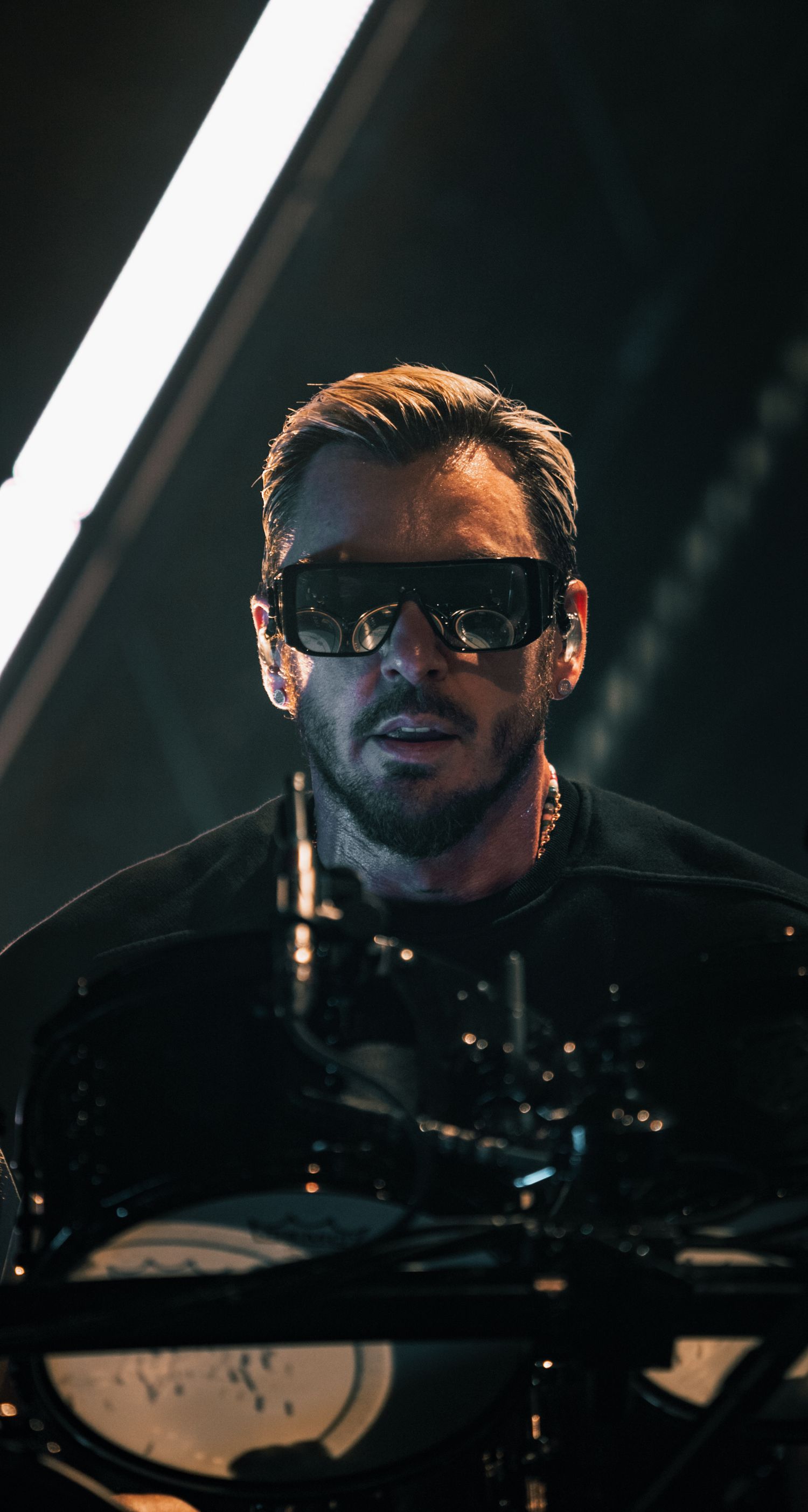A performer in dark sunglasses illuminated by stage lighting behind drum equipment.