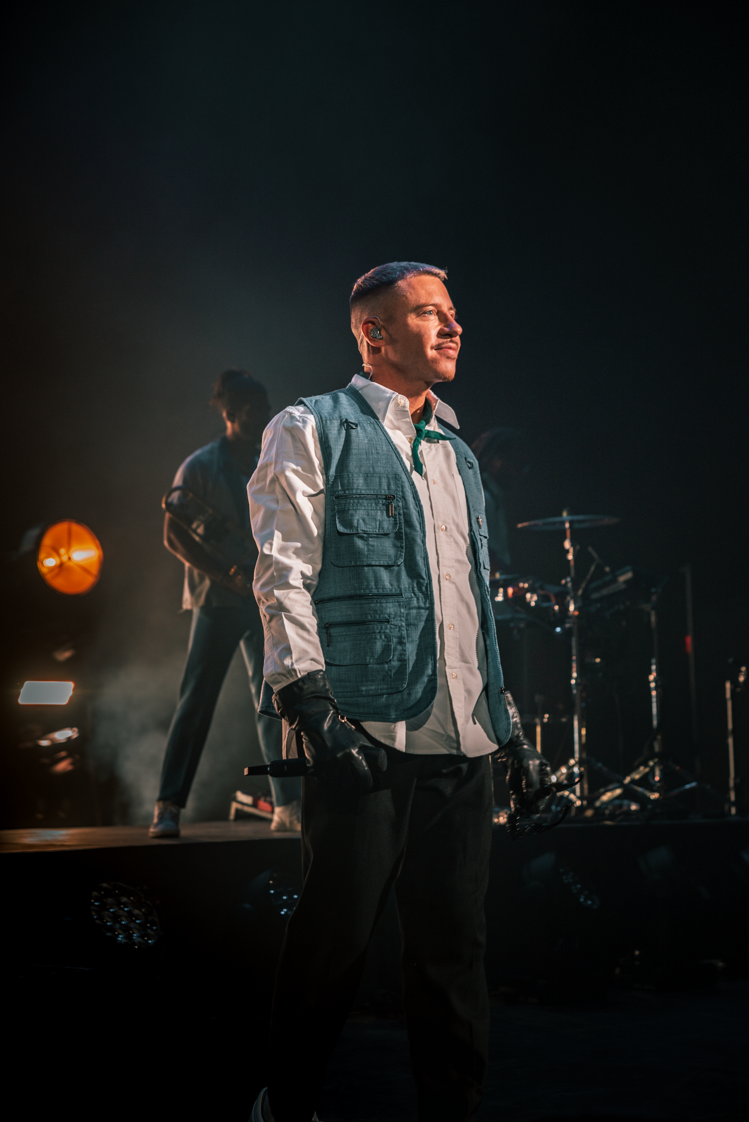 Concert performer in white vest illuminated against dark stage with orange lighting effects.