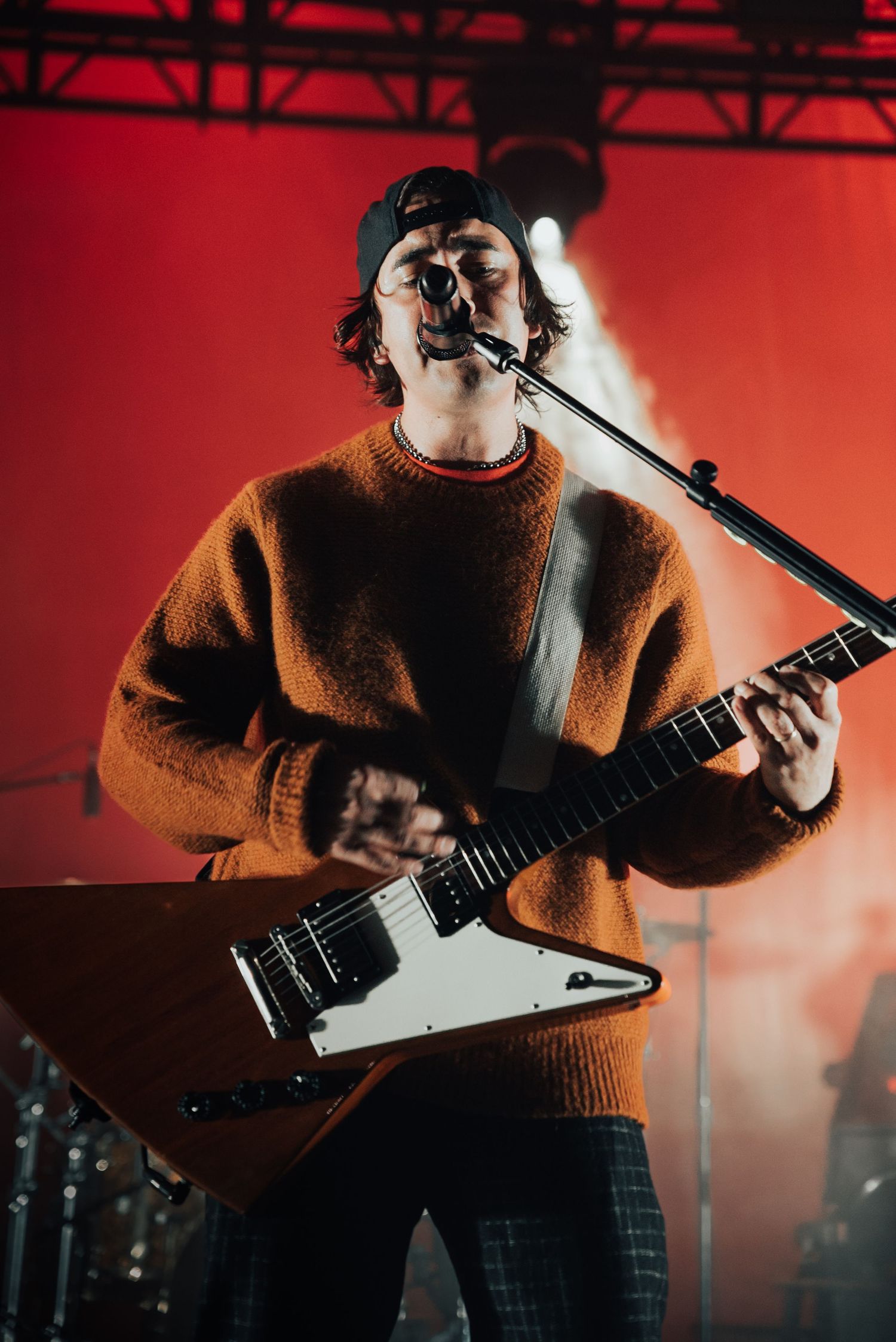 Guitarist in cozy sweater performs passionately with angular electric guitar under red stage lighting.