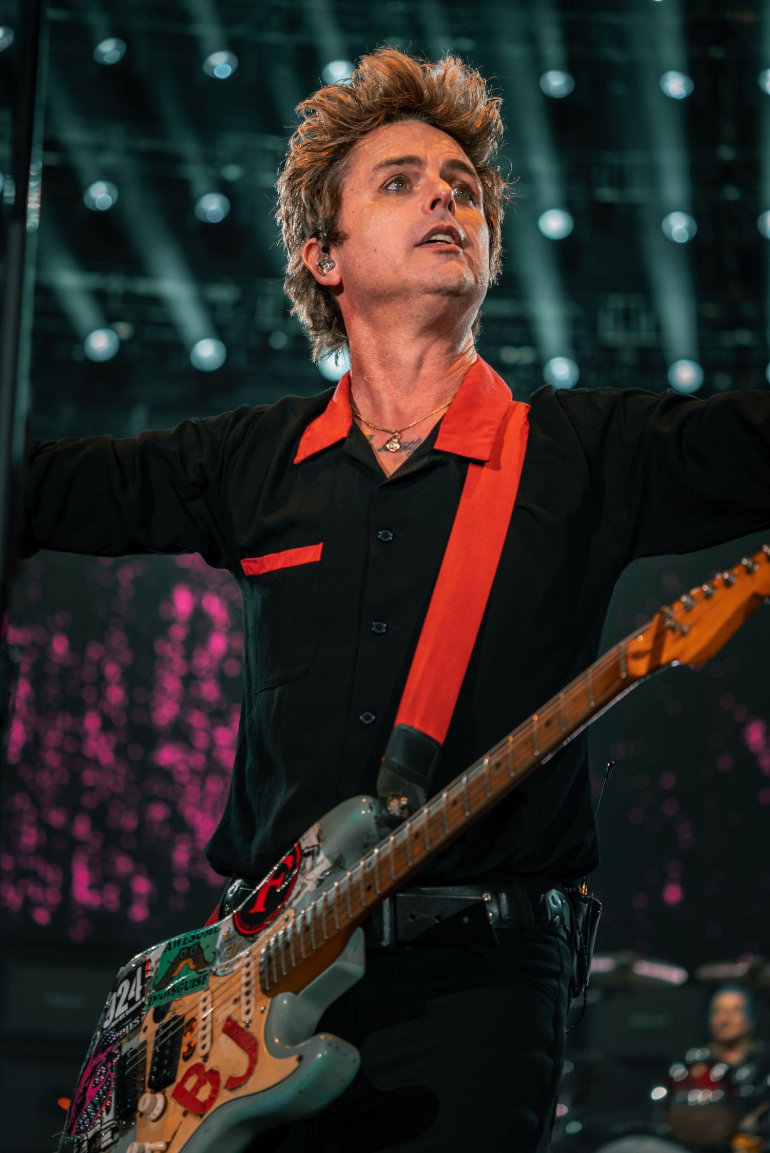 A performer in black with red accents plays a decorated guitar against a backdrop of stage lighting.