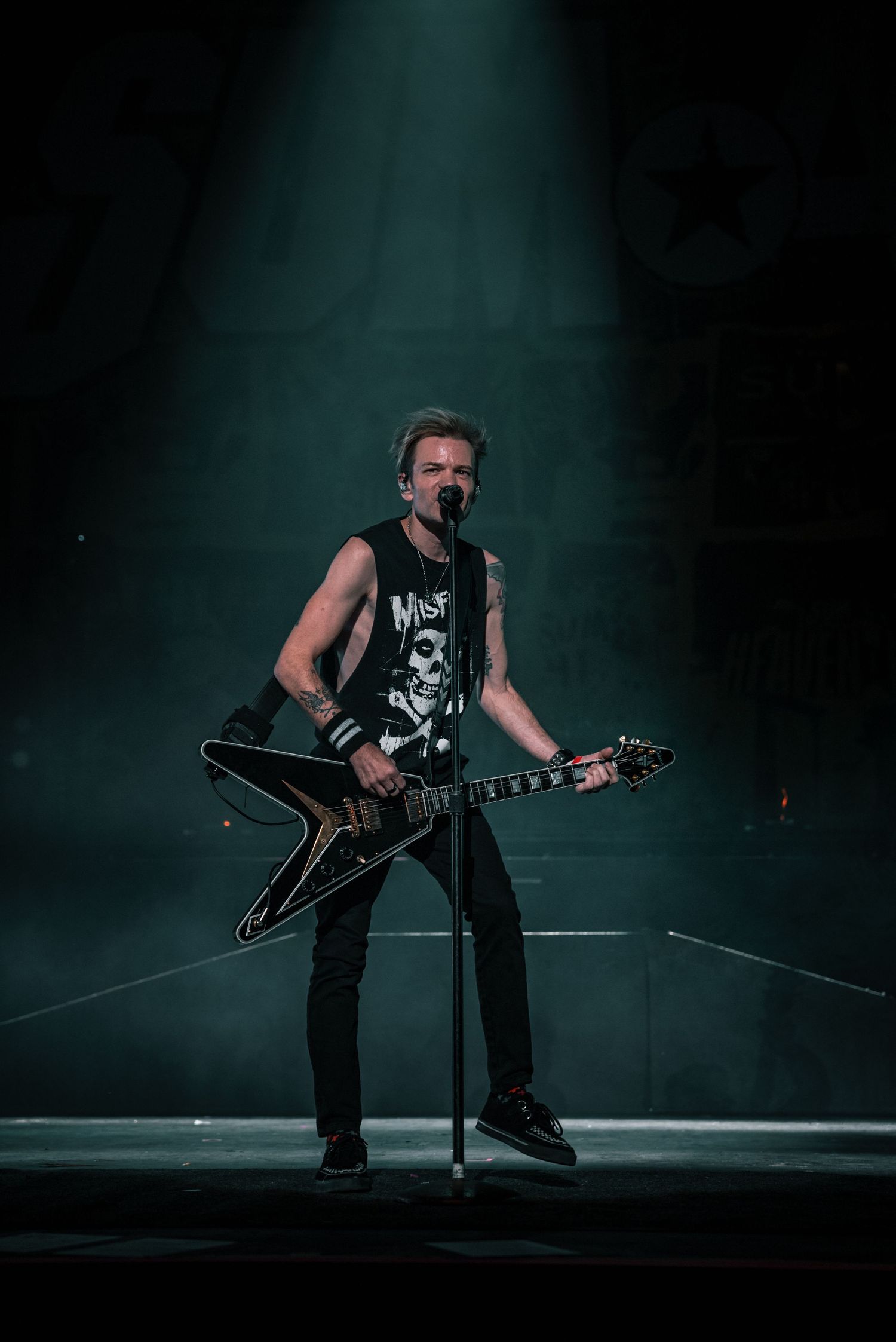 Guitarist in dark clothing performs dramatic pose while playing on dimly lit stage.