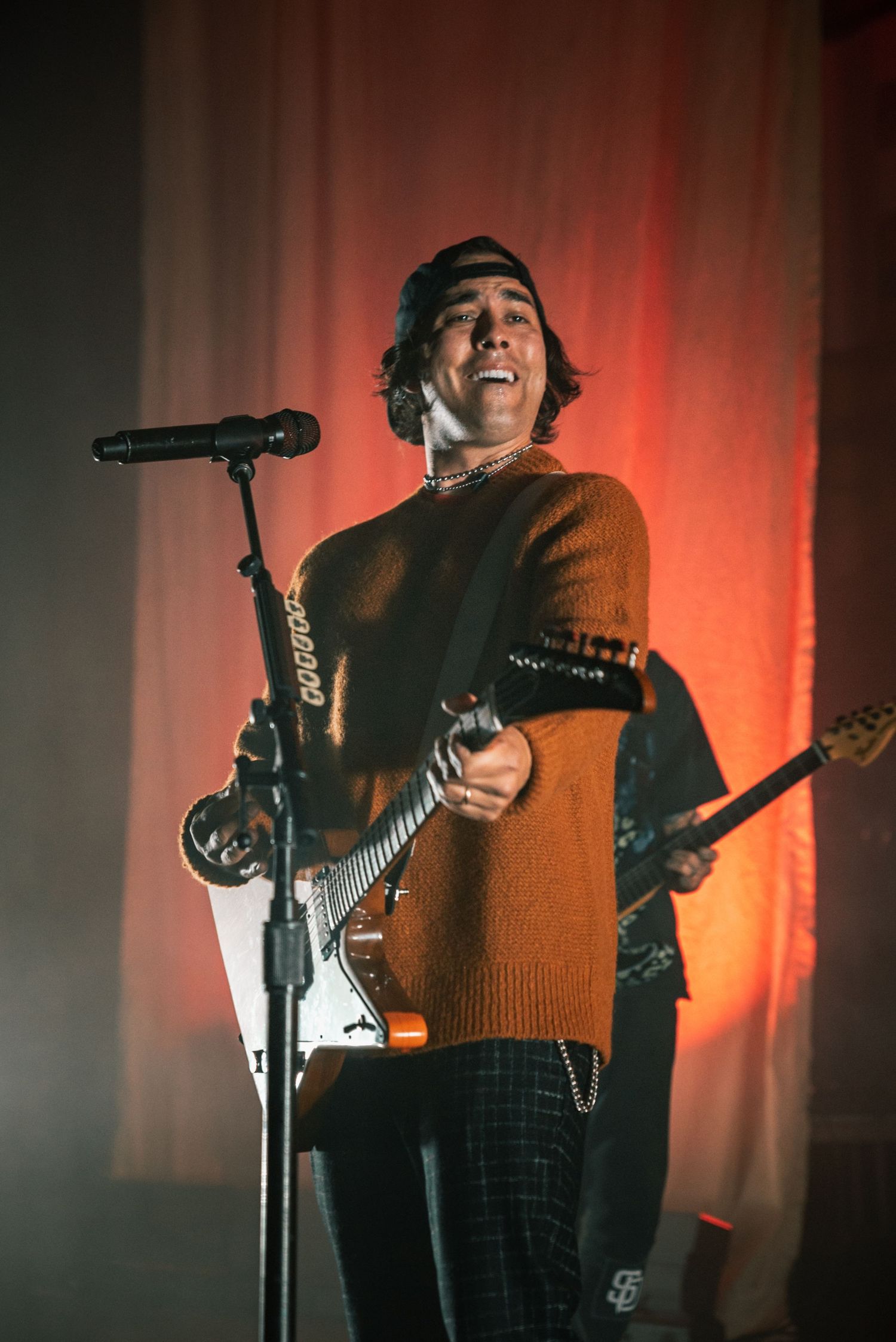 Musician in brown sweater performs with electric guitar under dramatic red stage lighting.