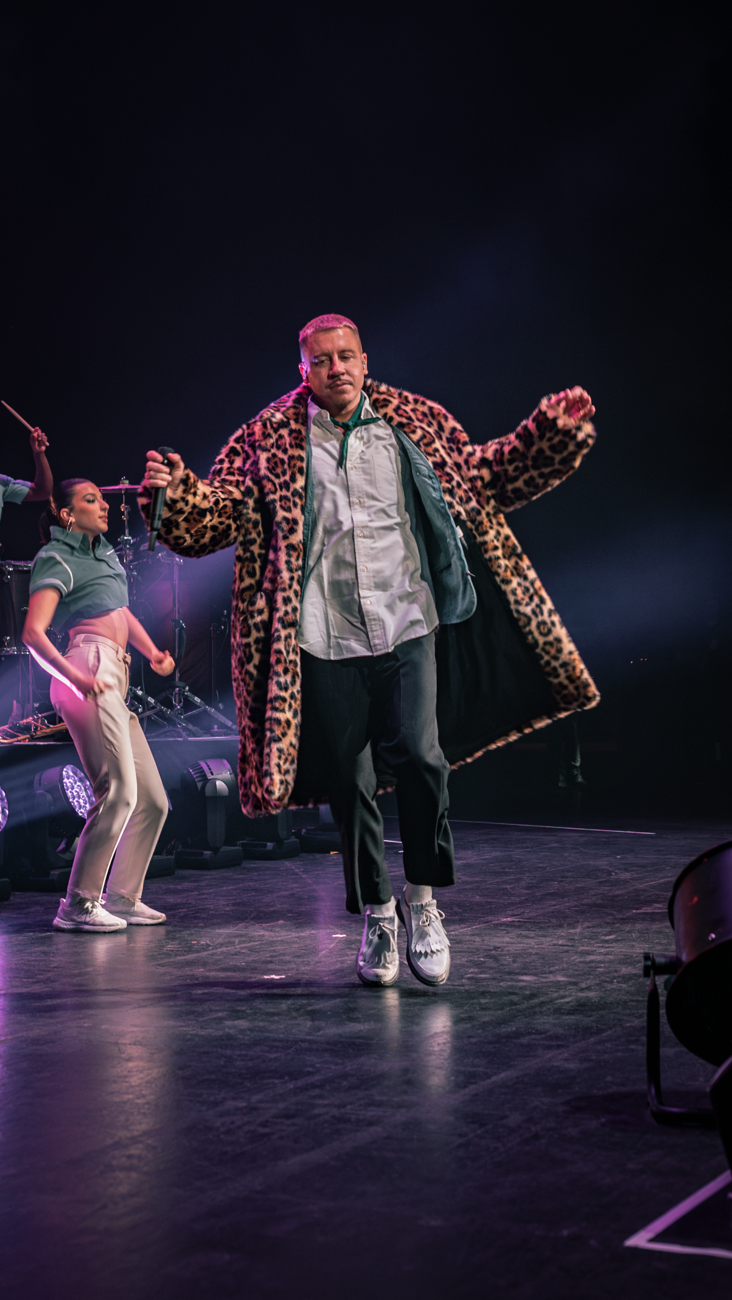 Performer in leopard print coat dances alongside backup dancers on a well-lit concert stage.
