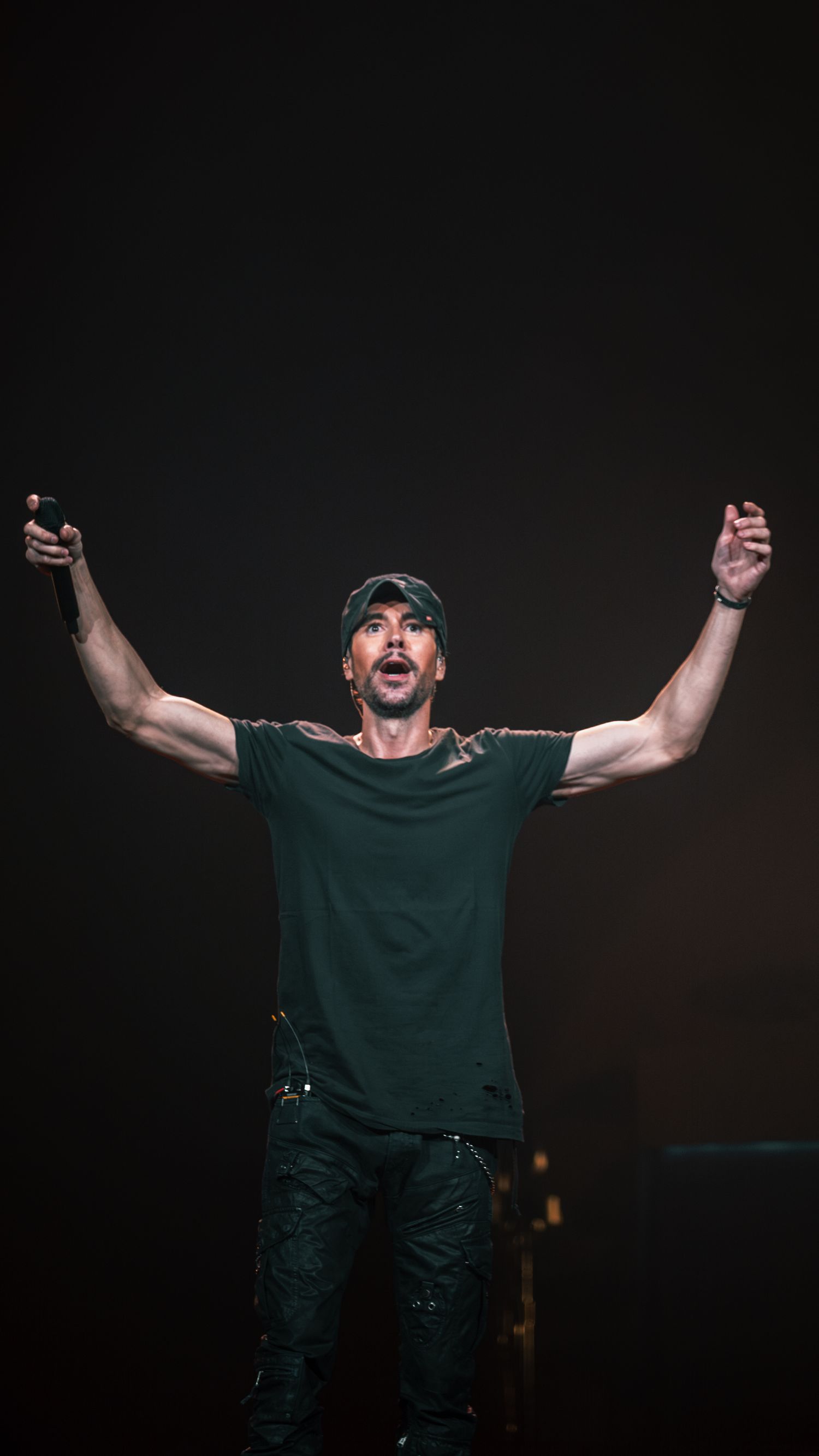 Performer in green shirt passionately raises arms during dramatic concert performance on dark stage.