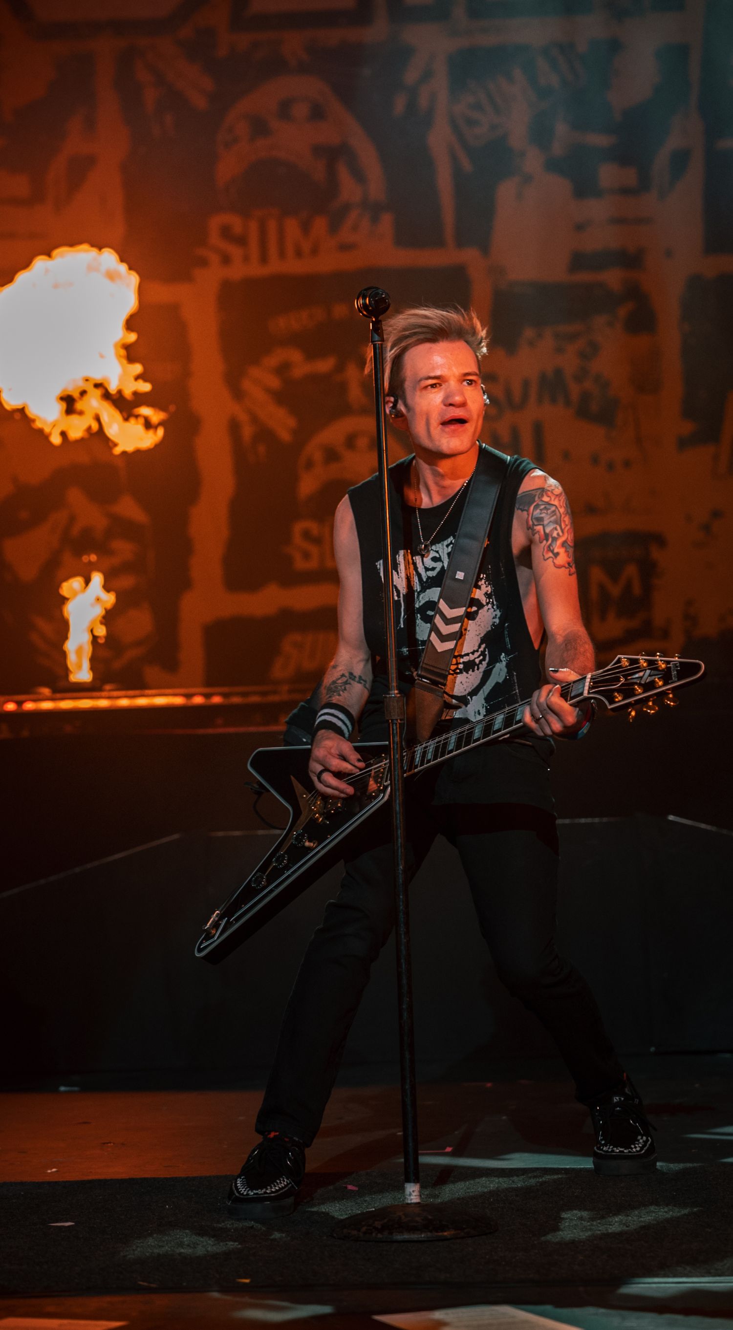 Rock performer plays V-shaped guitar with fiery pyrotechnics and graffiti wall backdrop.
