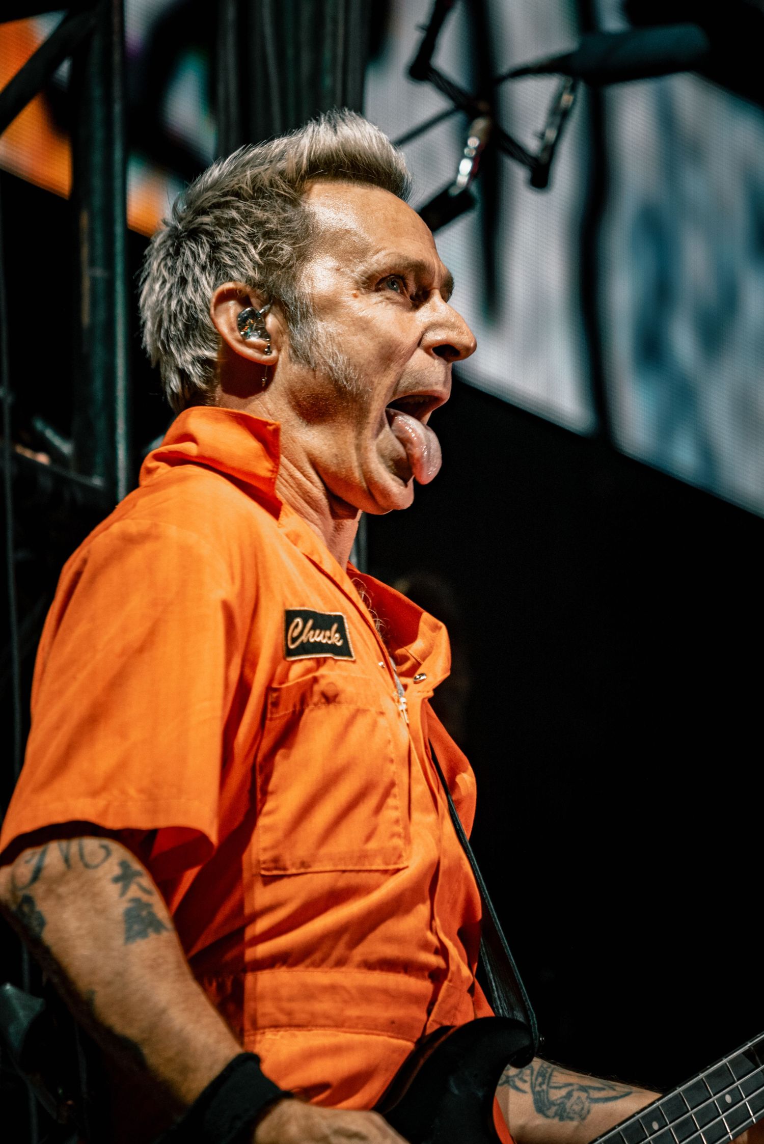 A musician in orange workwear performs energetically on stage with visible tattoos and in-ear monitors.