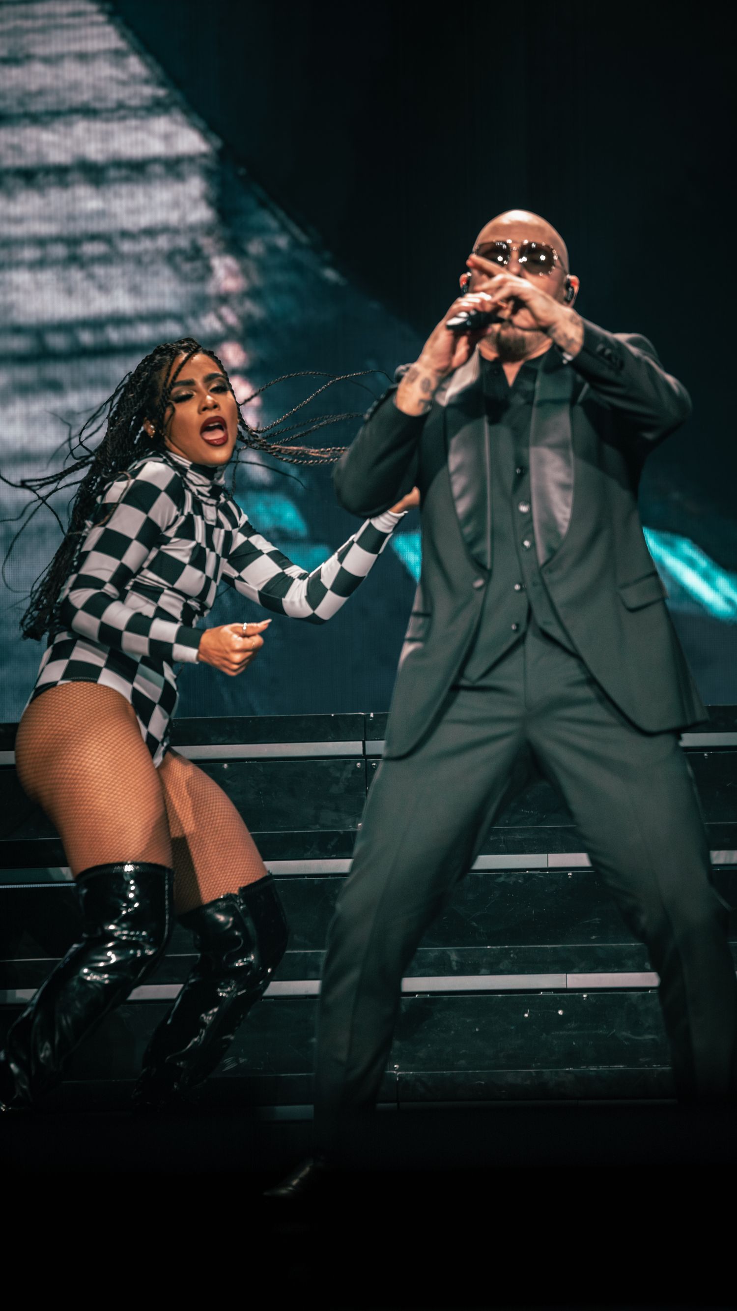 Energetic stage performance featuring dancers in checkered outfits alongside suited performer on elevated platform.