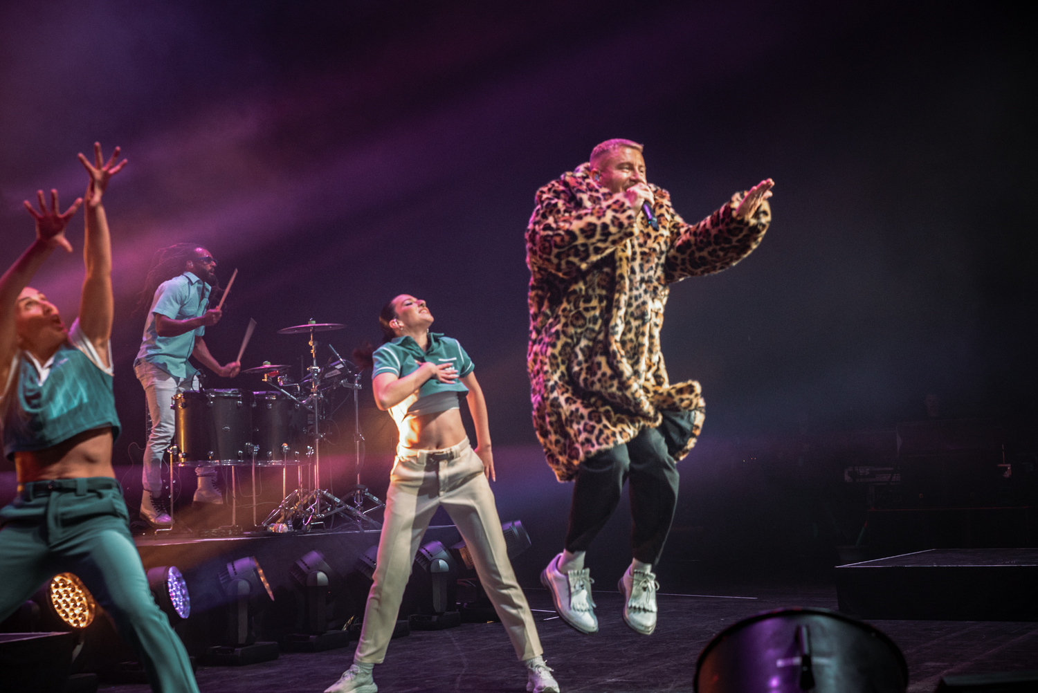 High-energy performance on stage with dancers and performer in leopard print coat mid-jump.