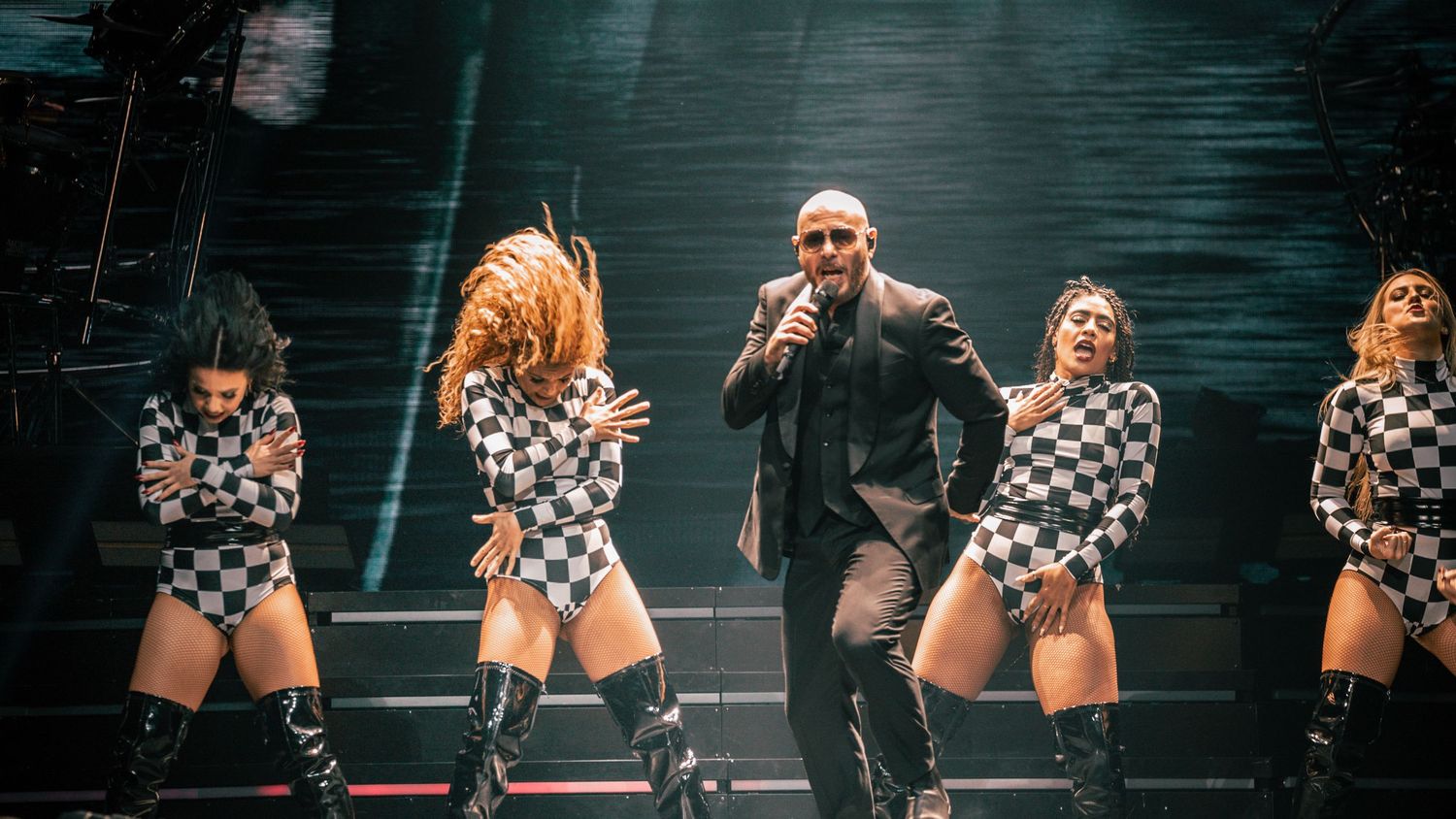 Performers deliver high-energy dance routine in black and white checkered costumes on concert stage.