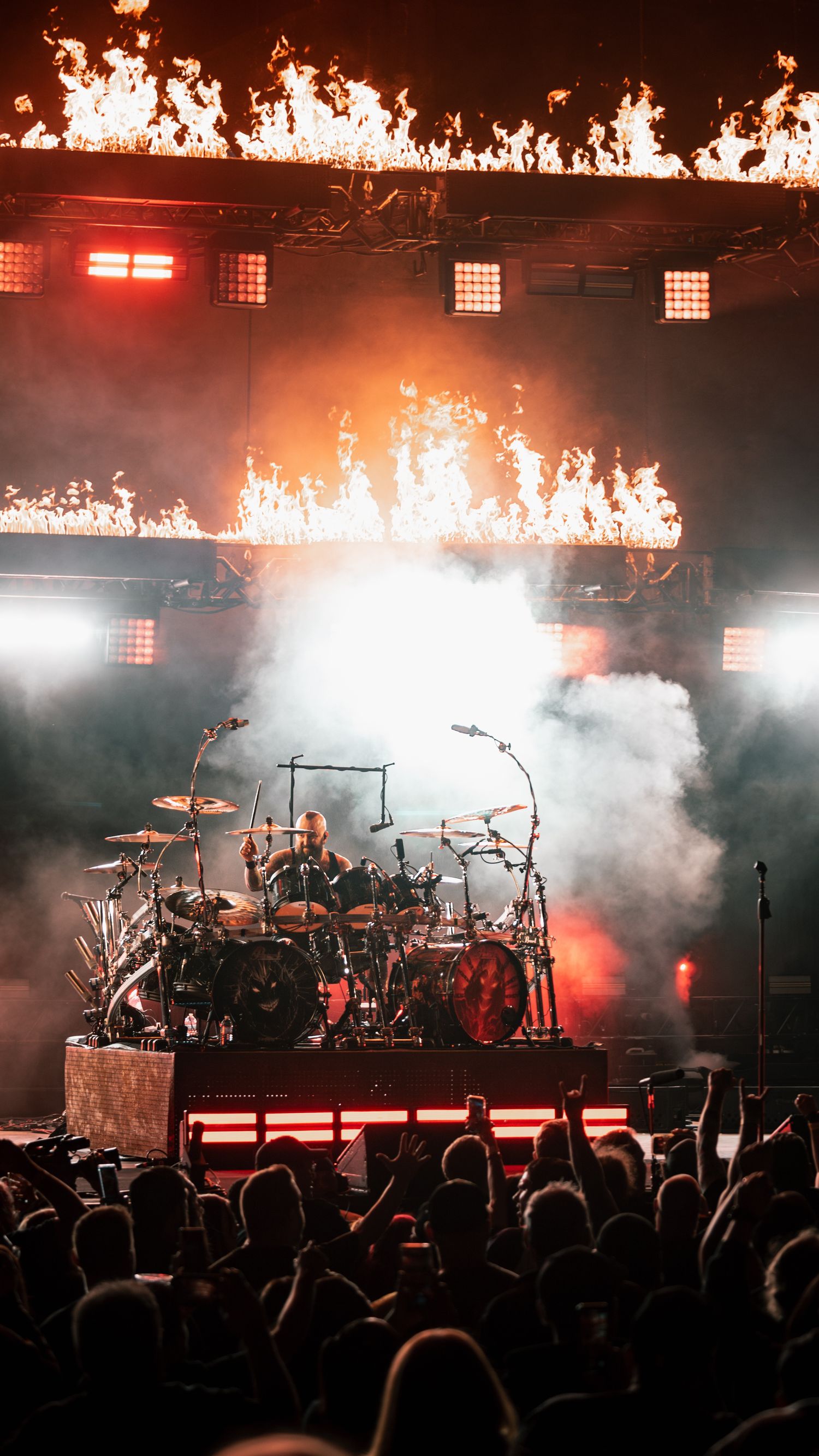 Concert stage setup with fire effects, drum kit and equipment silhouetted against bright red lighting.