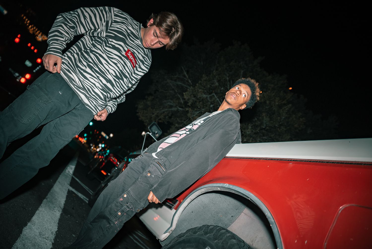 Street fashion photo with two people posing against a red car at night.