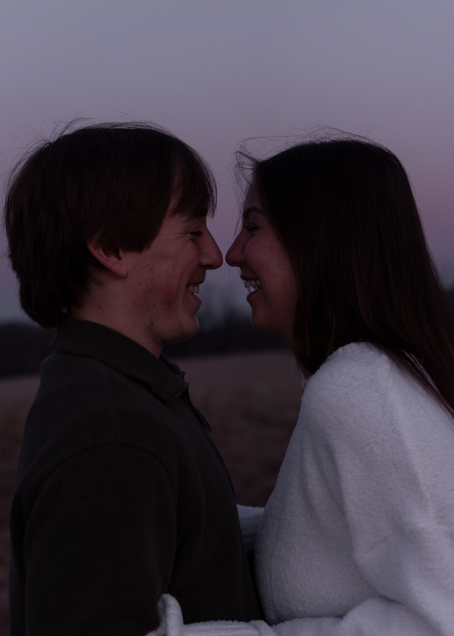 Laughing couple facing each other at twilight in an open field during a romantic engagement session.