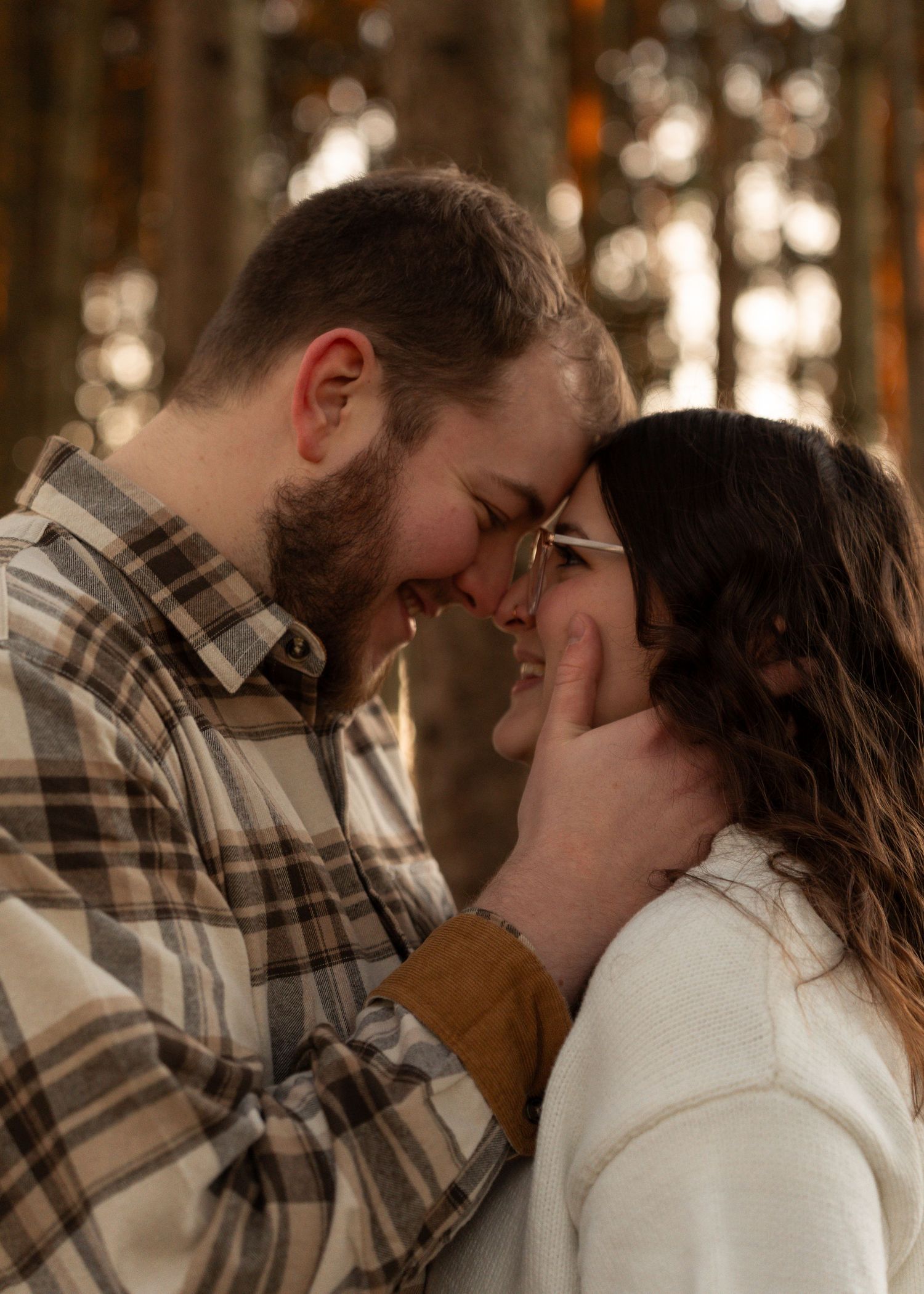 Couple laughing and touching foreheads tenderly in a sun-dappled forest setting.