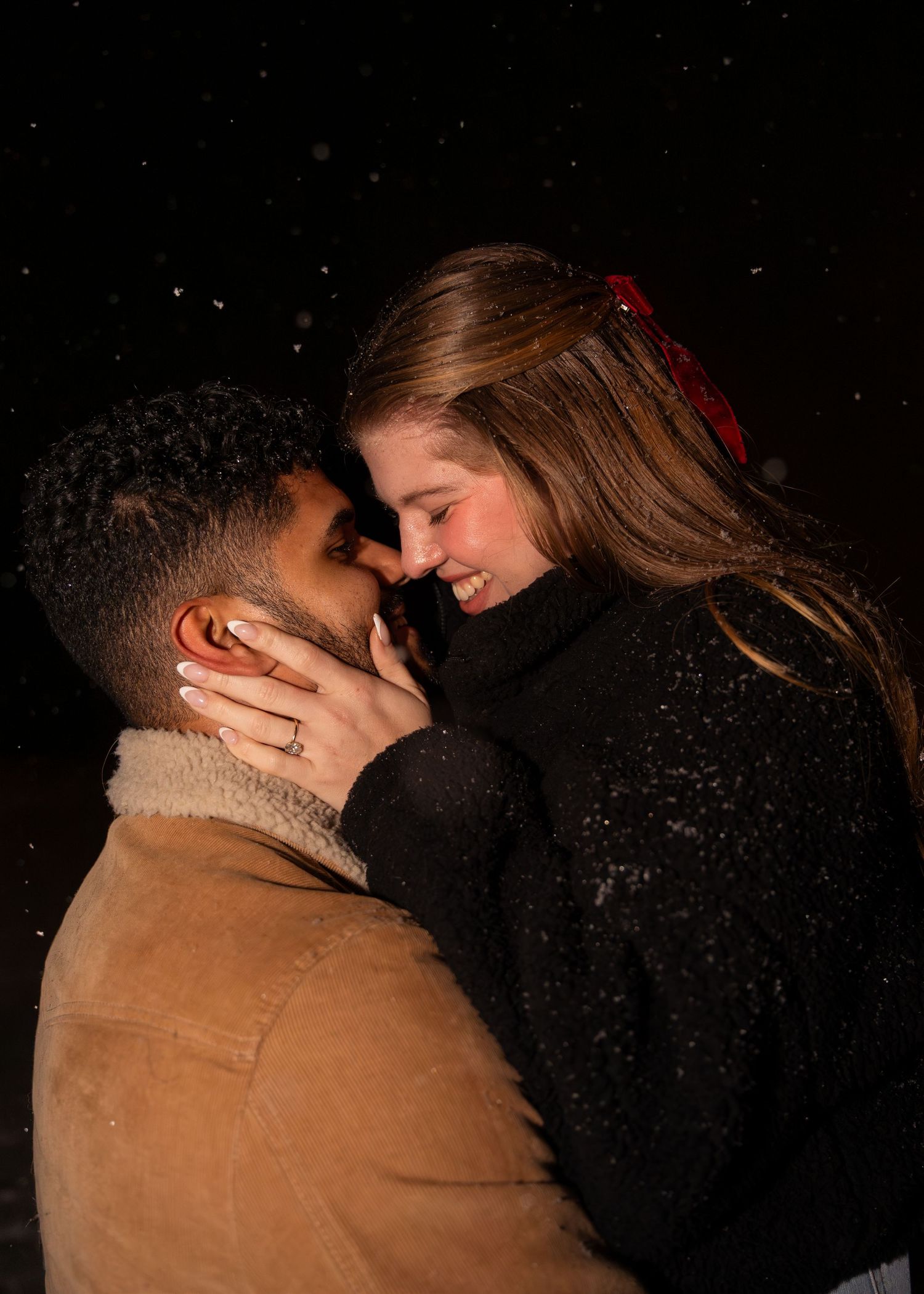 Couple sharing a romantic moment in the snow at night, woman wearing a red bow and engagement ring.