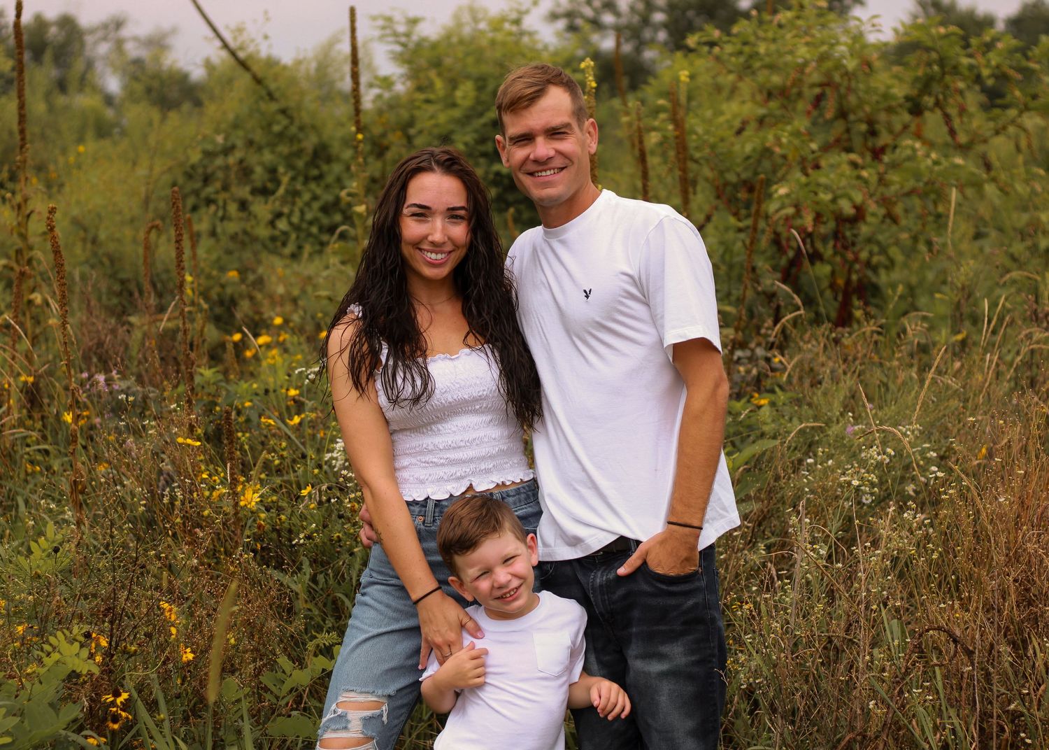 Happy family portrait of parents and young child smiling together in a wildflower meadow setting.
