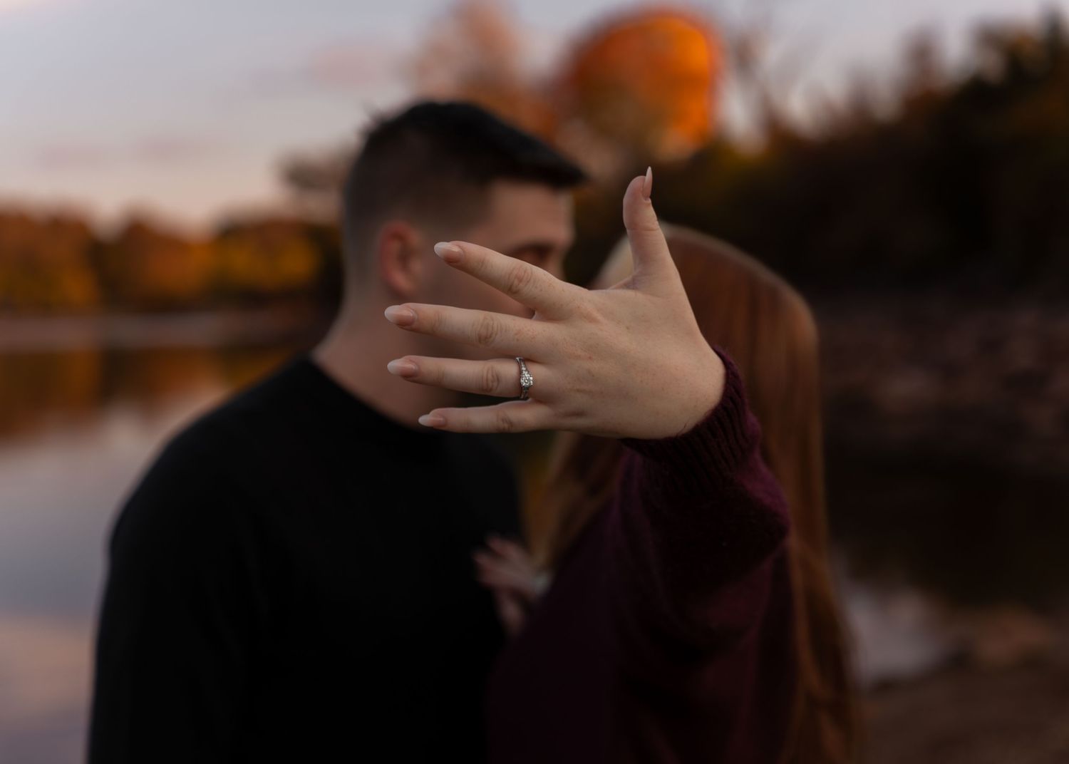 Couple sharing a kiss outdoors while woman displays engagement ring to camera near autumn lake.